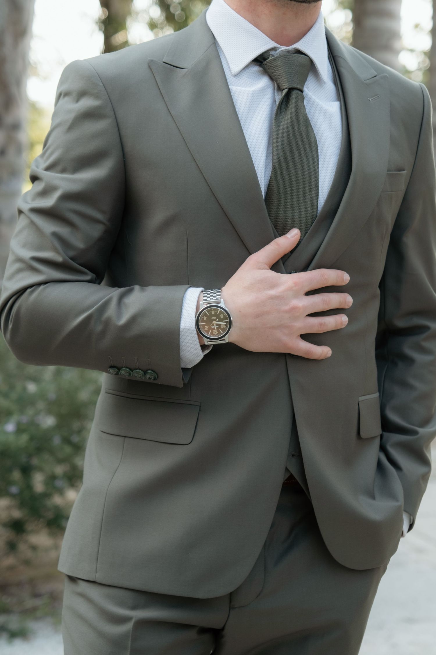 Man in olive gray three-piece suit with matching tie and silver watch, hand placed on chest.