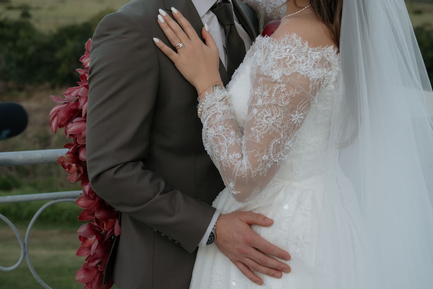Bride in lace wedding gown and groom in gray suit embrace during outdoor wedding ceremony.