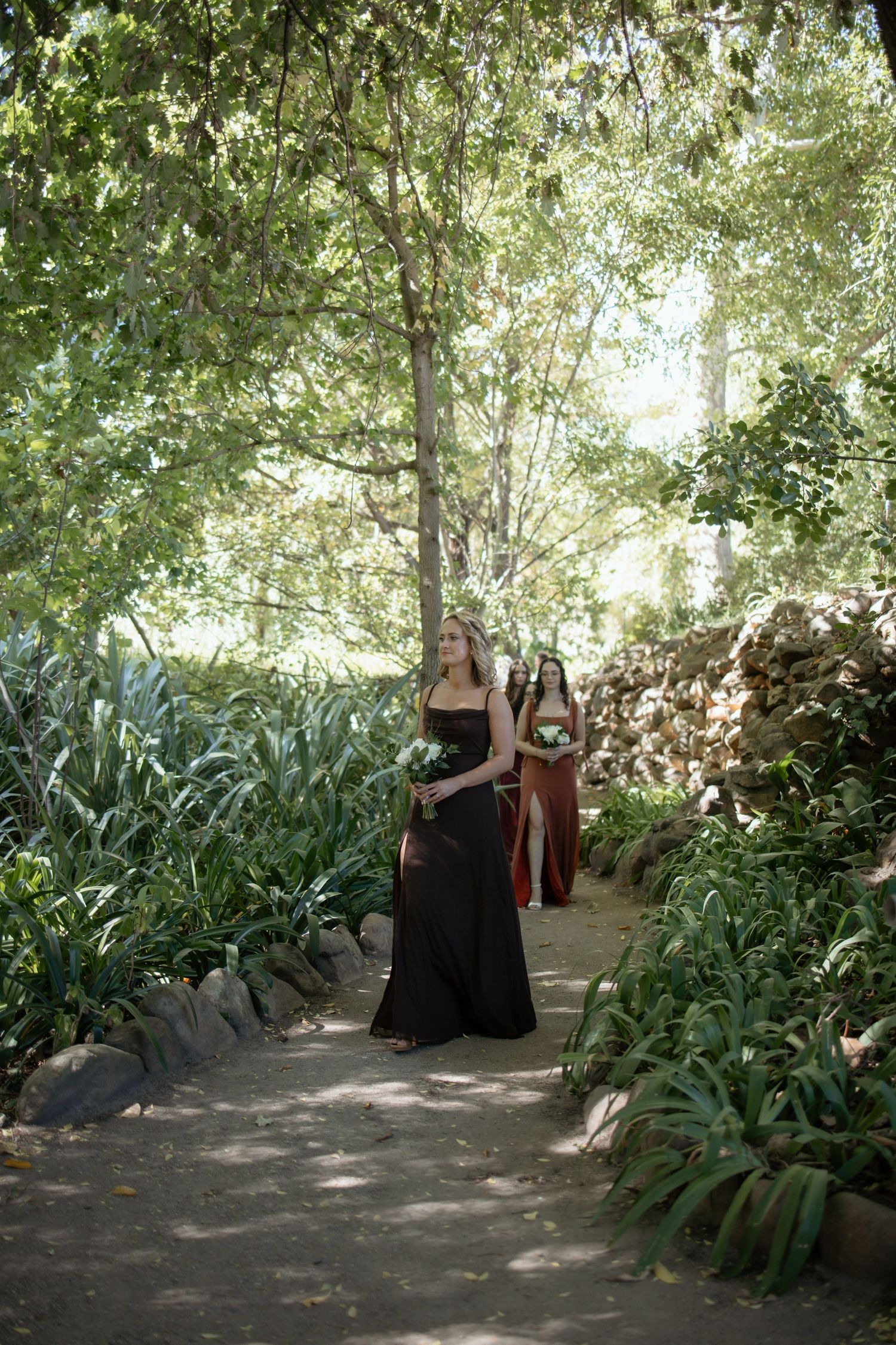 Bridesmaids in black and rust gowns walk down a lush garden path lined with tropical plants and trees.