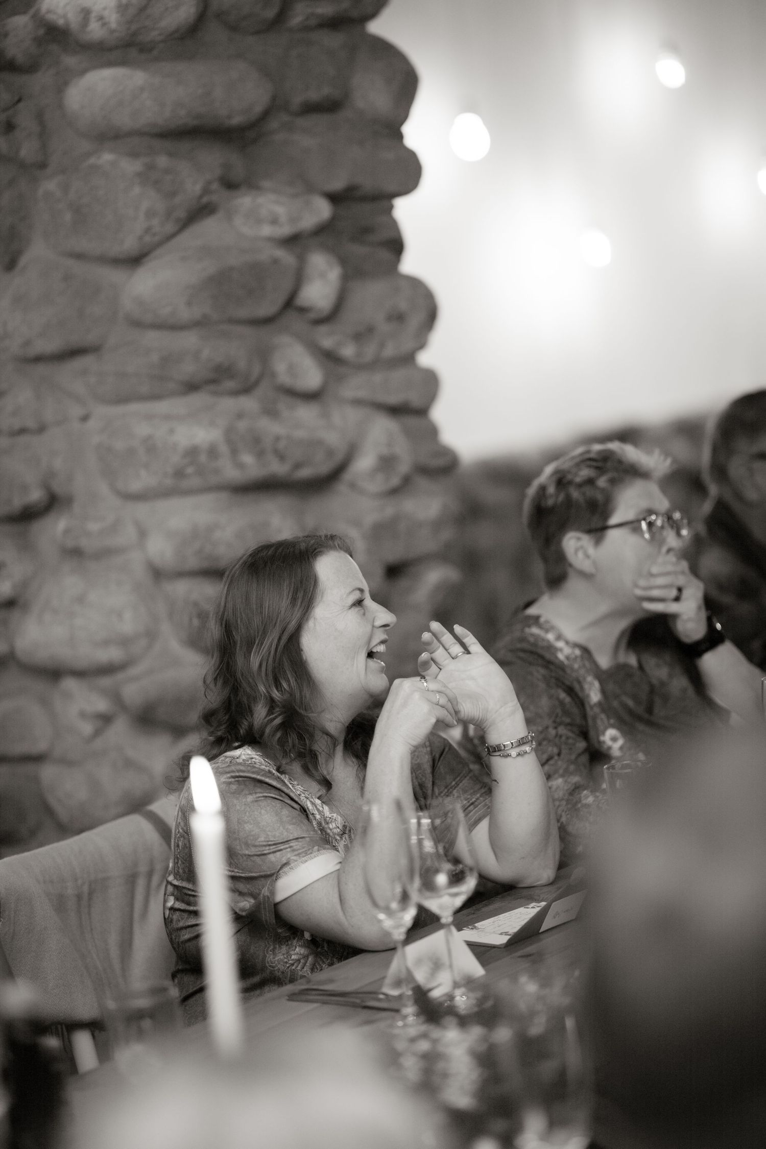Wedding guest laughs and claps during reception speeches beside stone wall with string lights above.