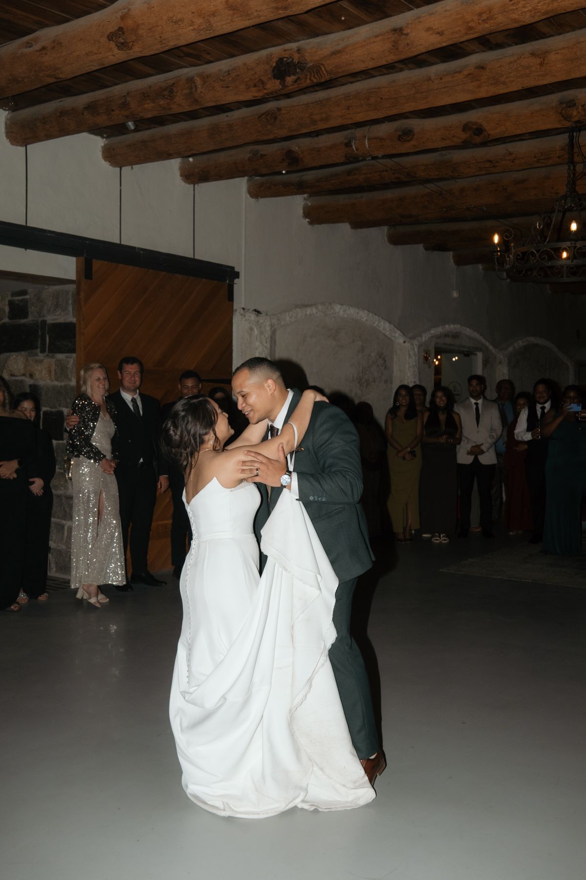 Newlyweds share their first dance on the floor of a rustic barn venue with guests watching.