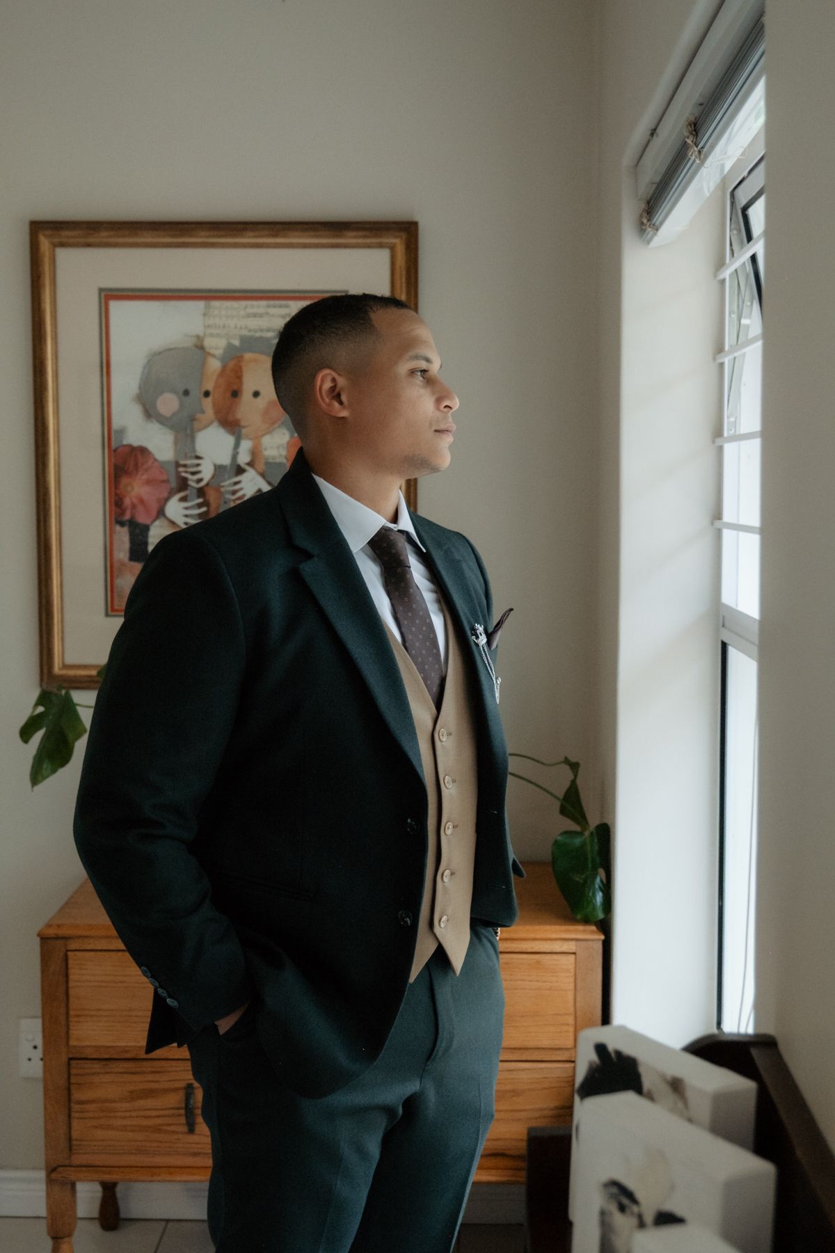 Dapper groom in dark suit with tan vest gazes thoughtfully out window during wedding day preparations.