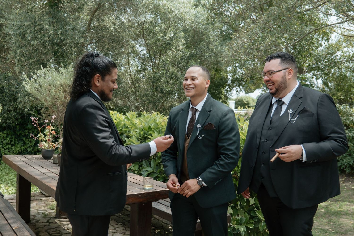 Three men in dark suits share a joyful conversation outdoors at a wedding, surrounded by lush greenery.