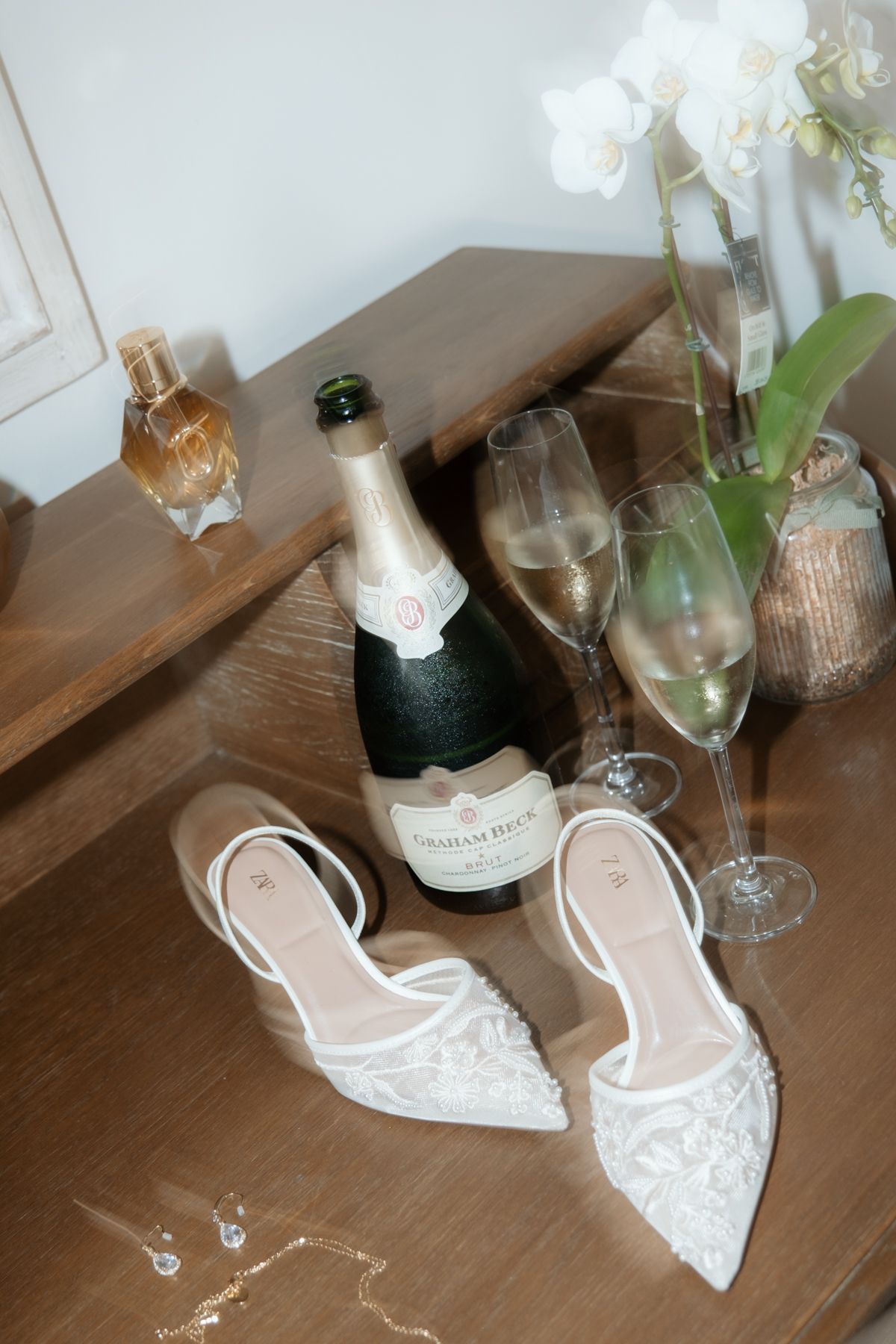 Bridal flat shoes, champagne bottle, glasses, and orchids arranged on wooden table.