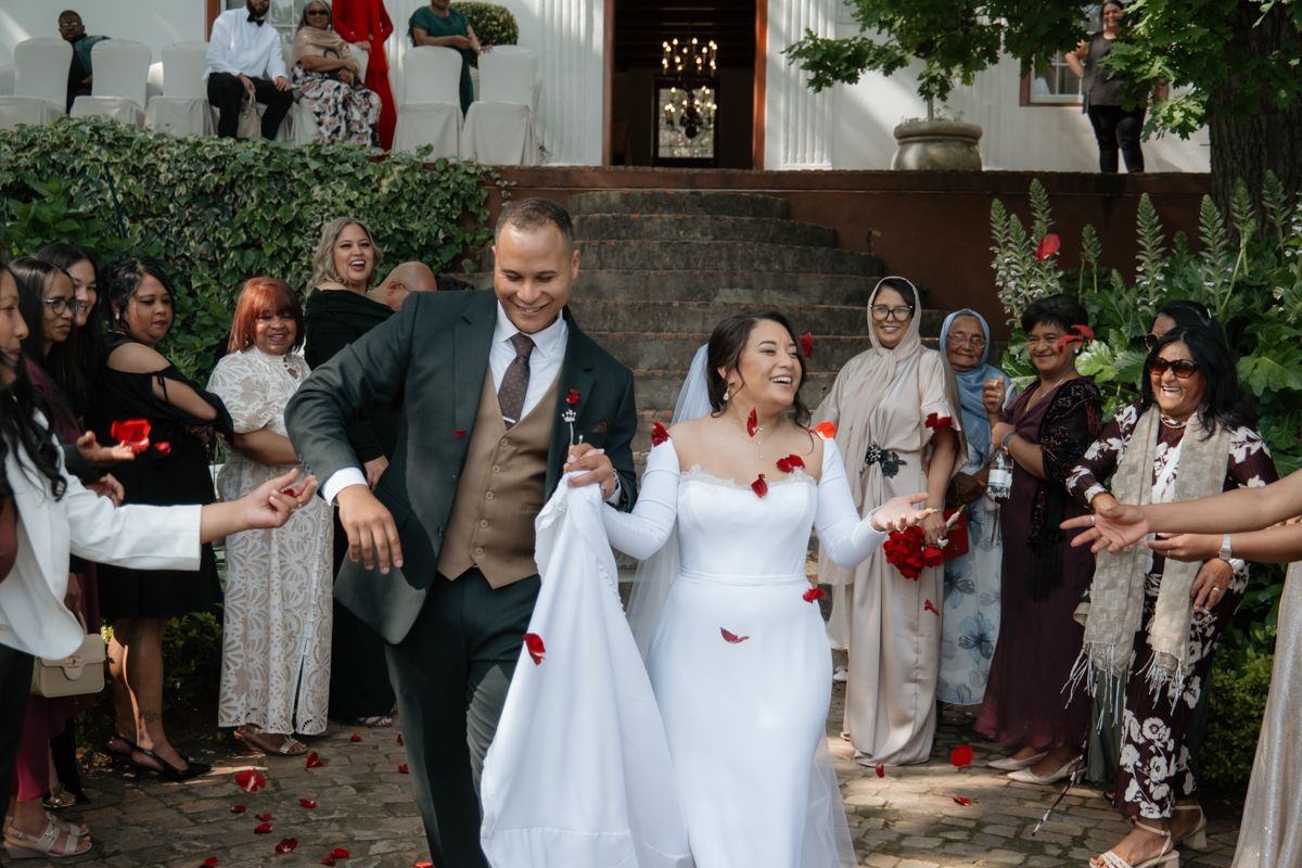 Joyful newlyweds walk through cheering guests tossing red rose petals after outdoor wedding ceremony.