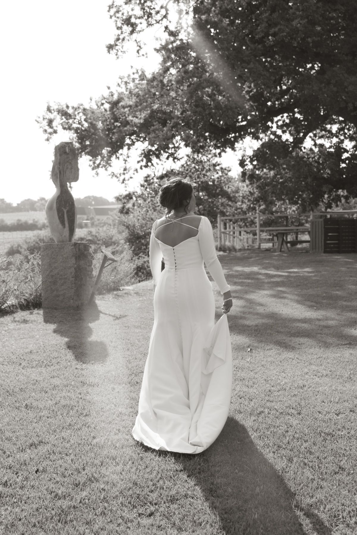 Bride in elegant white long-sleeve gown with open back stands on garden path near sculpture in black and white photo.