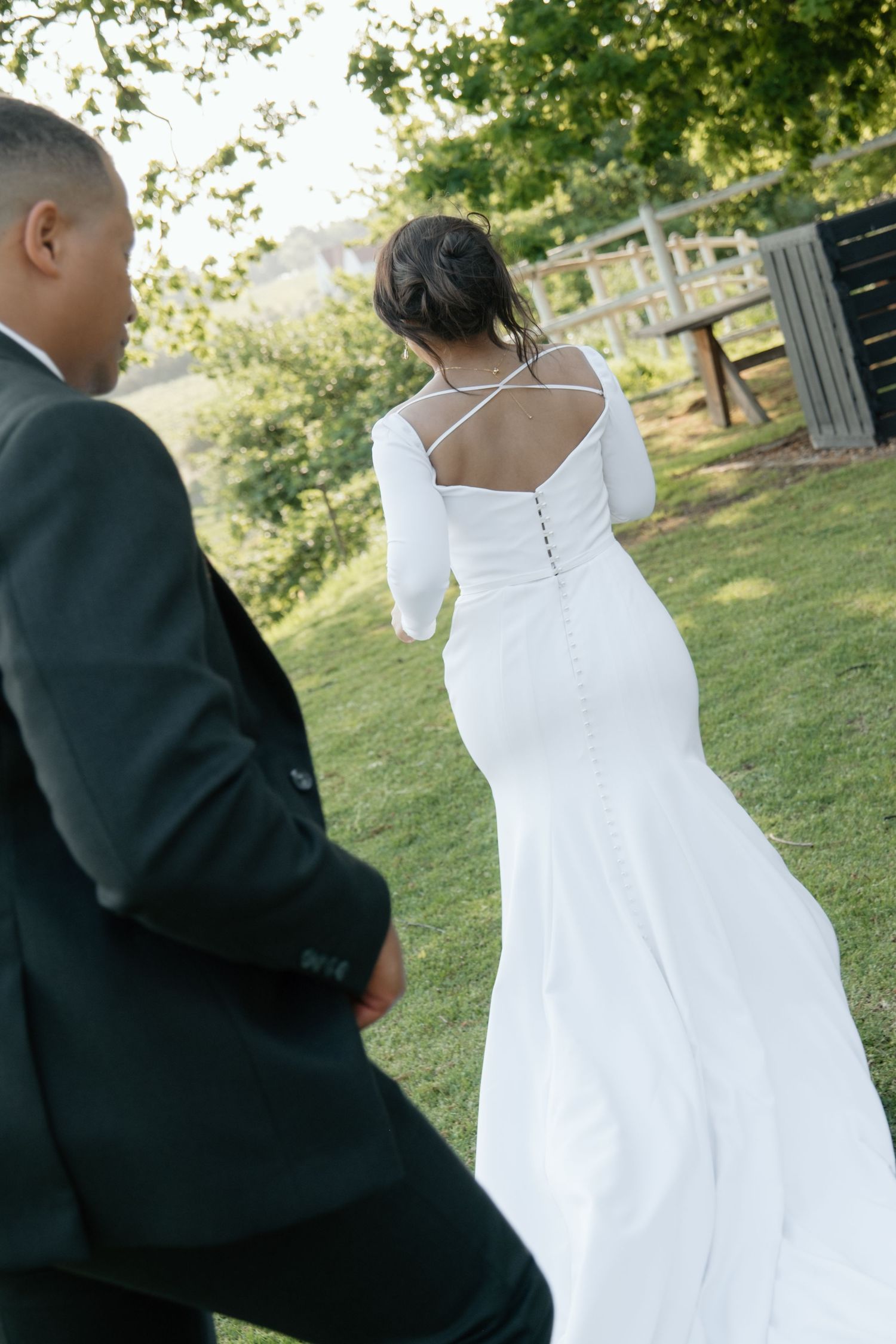 Bride in elegant backless white gown with button detail walks with groom through lush garden.