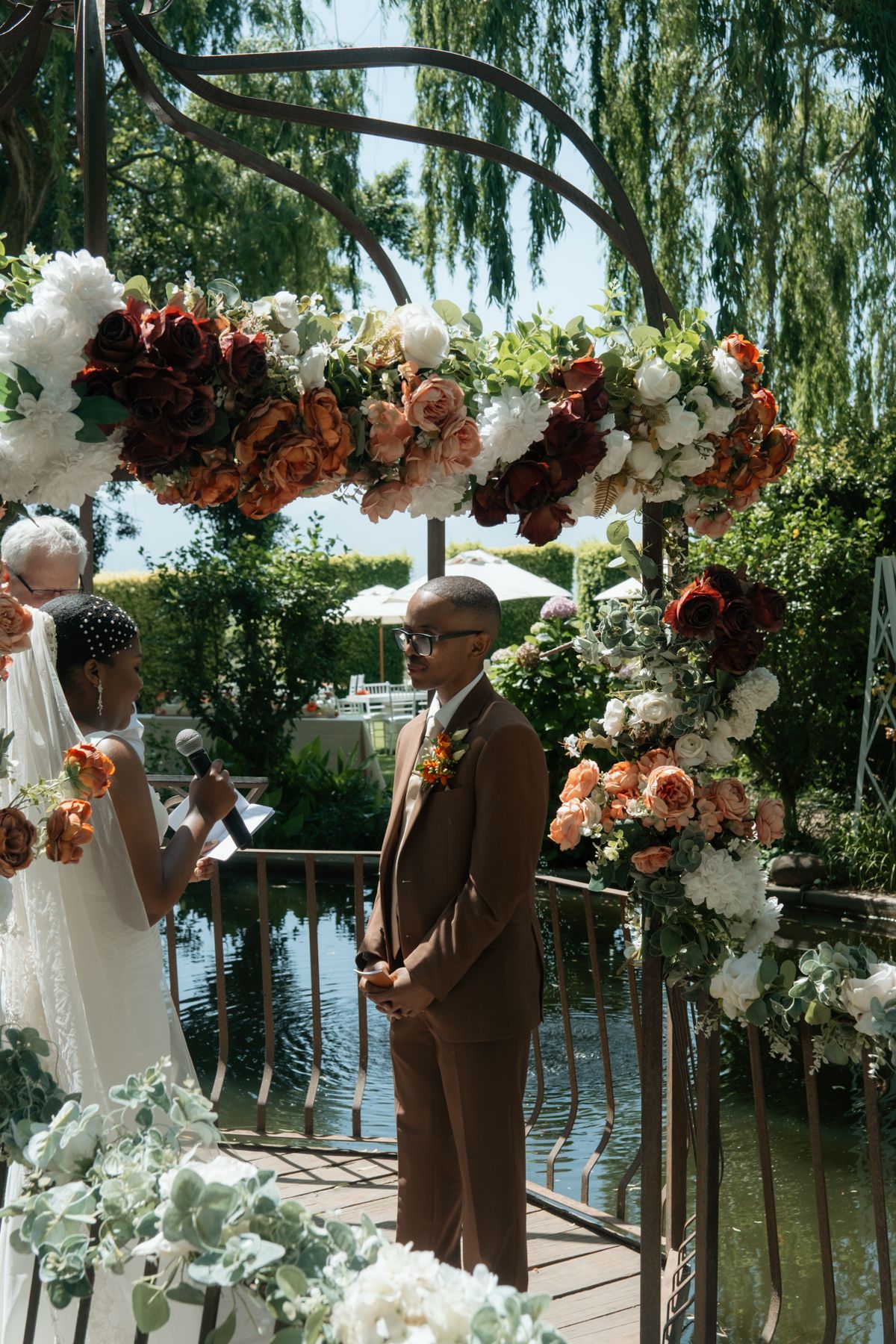 Groom in brown suit stands under colorful floral arch with bride at outdoor wedding.