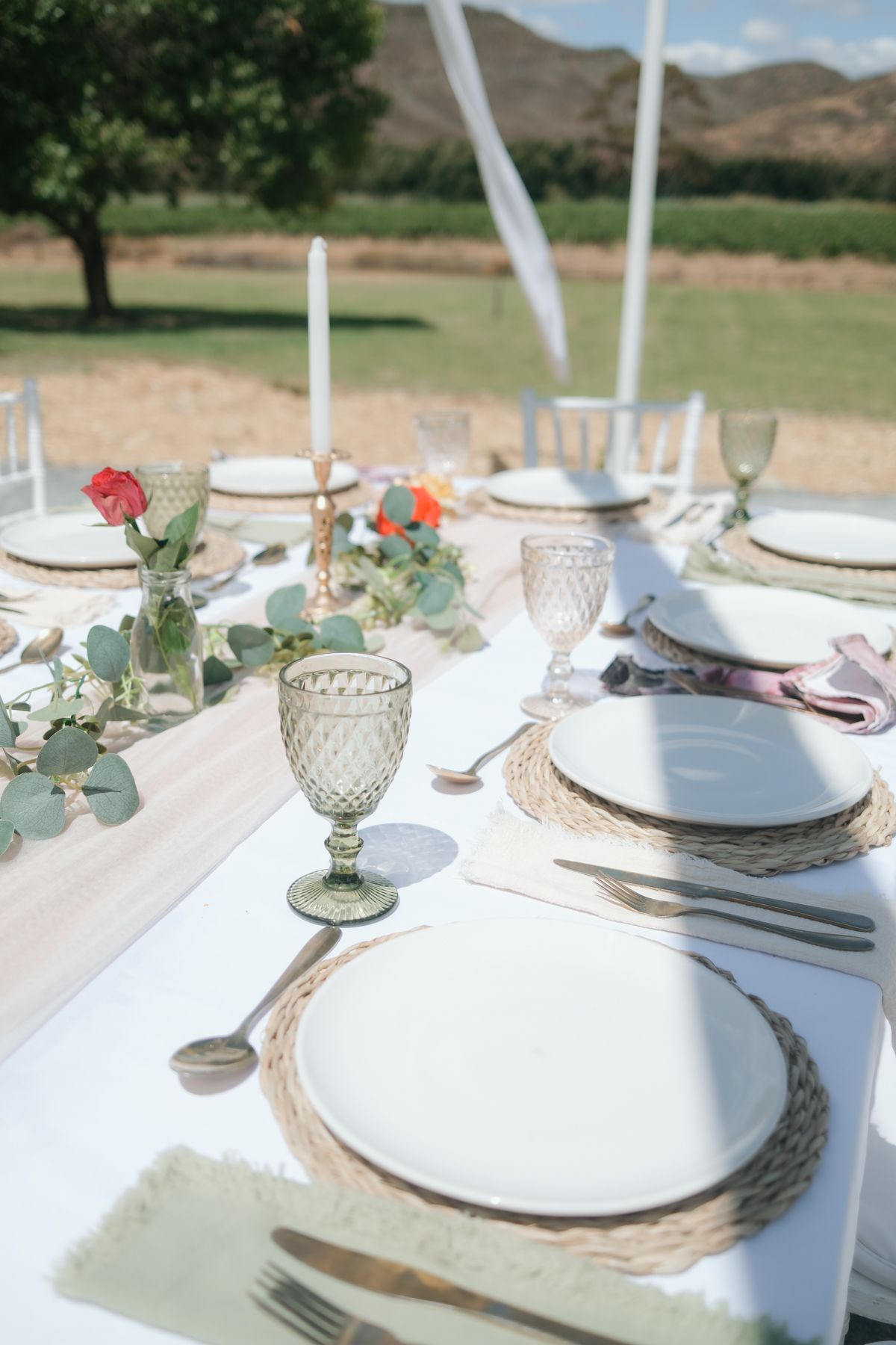 Elegant outdoor wedding table setting with woven chargers, green glassware, candles, and floral centerpiece.