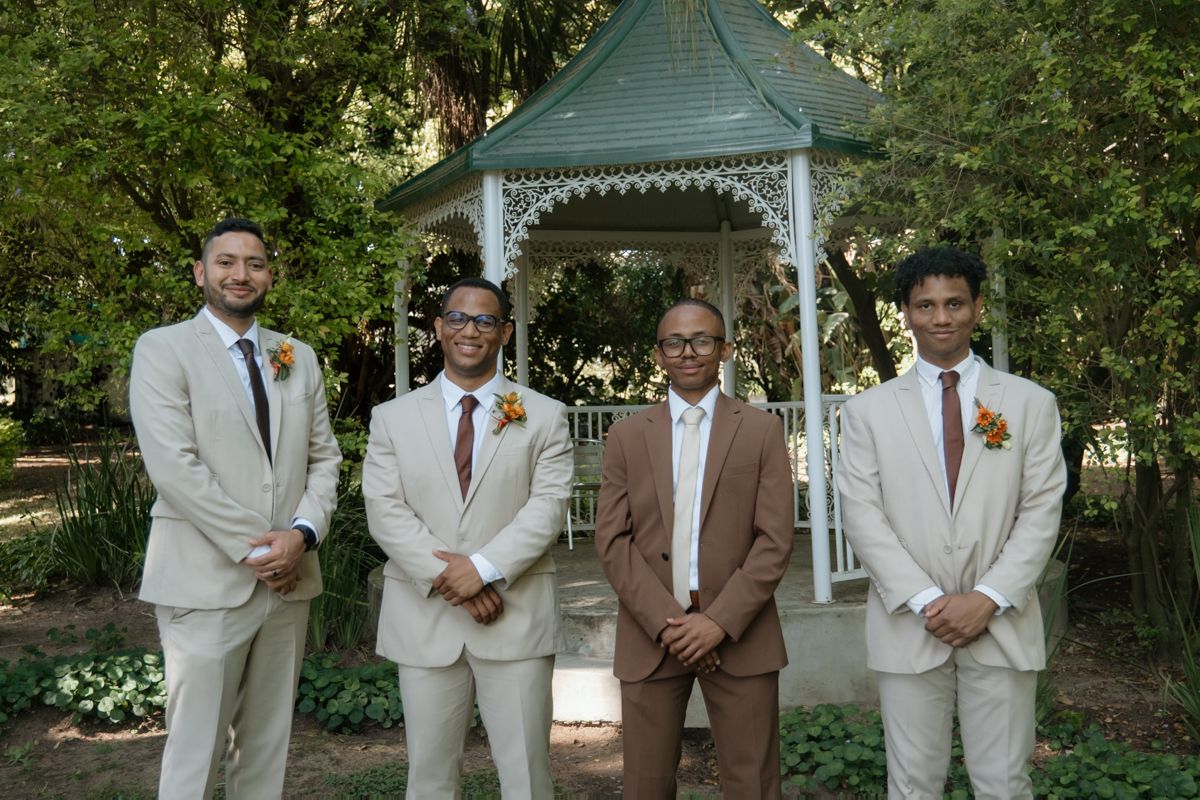 Four groomsmen in beige and brown suits pose together in front of white garden gazebo.