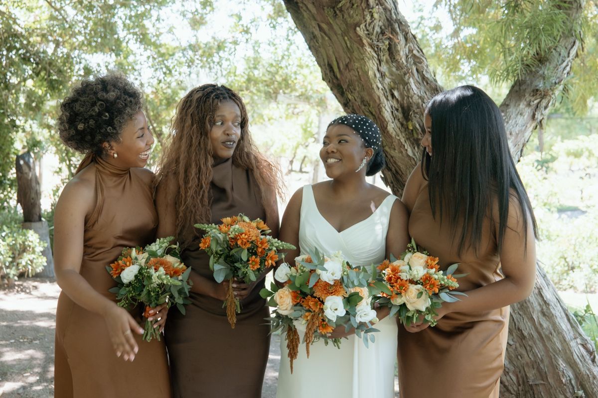 Bride in white gown with three bridesmaids in brown dresses holding orange and white bouquets outdoors.