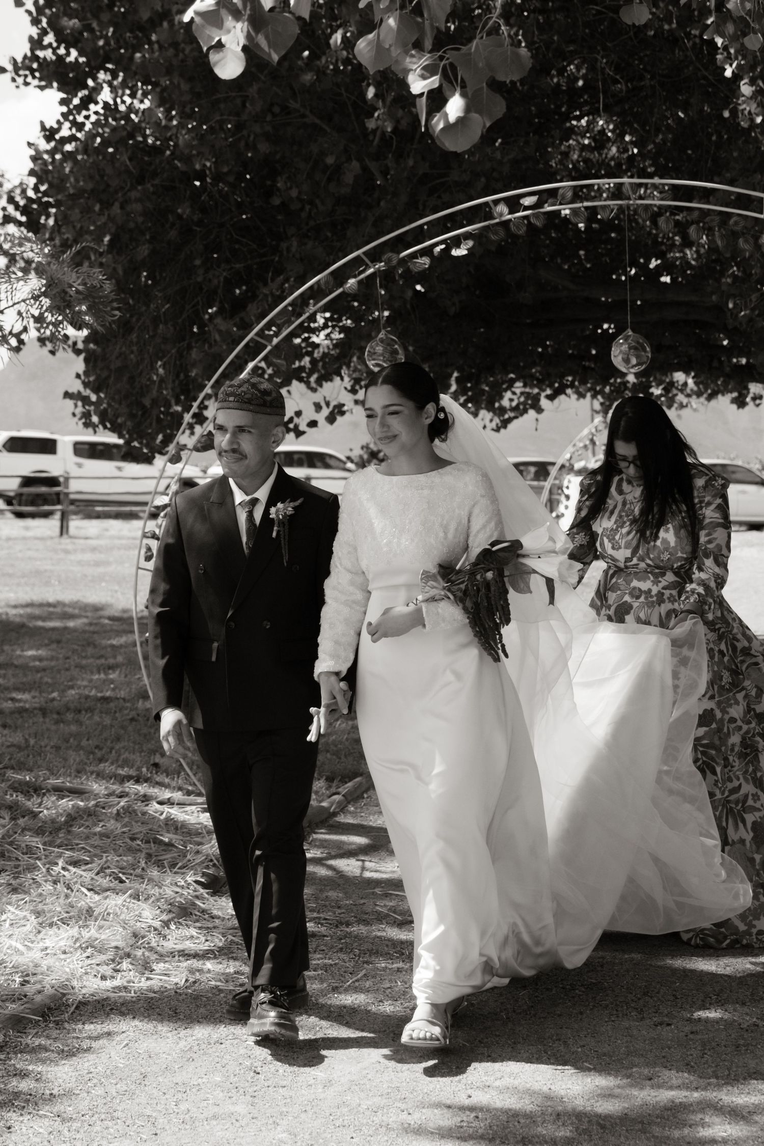 Bride in white gown walks with groom in black suit through garden arch in black and white wedding photo.