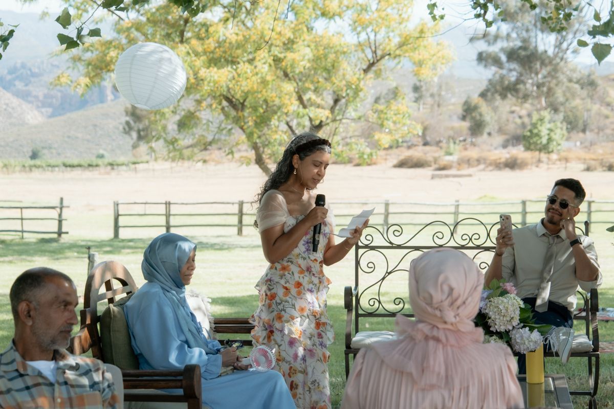 Woman in floral dress giving a speech at an outdoor wedding with mountain backdrop and paper lanterns.