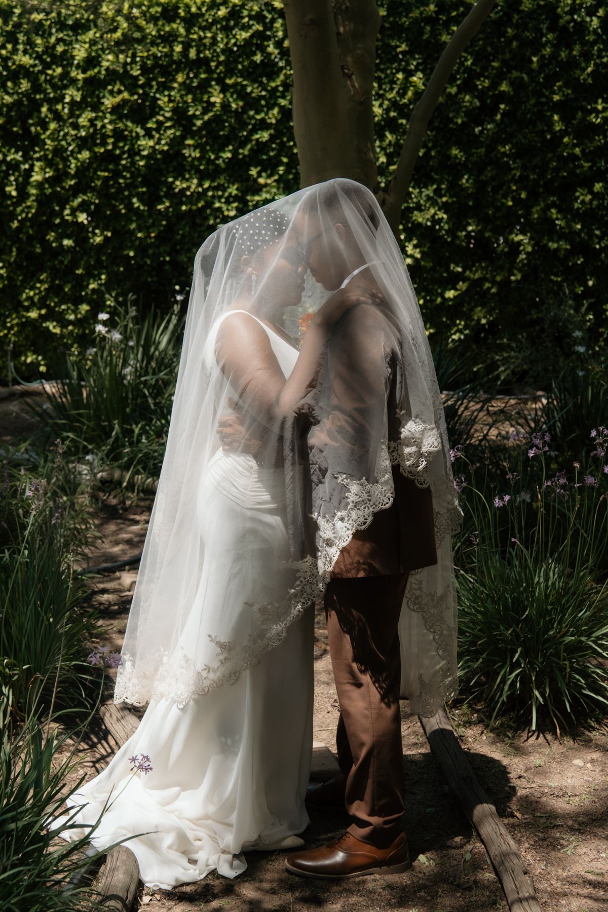 Bride and groom share romantic kiss hidden beneath lace wedding veil in sunlit garden setting.