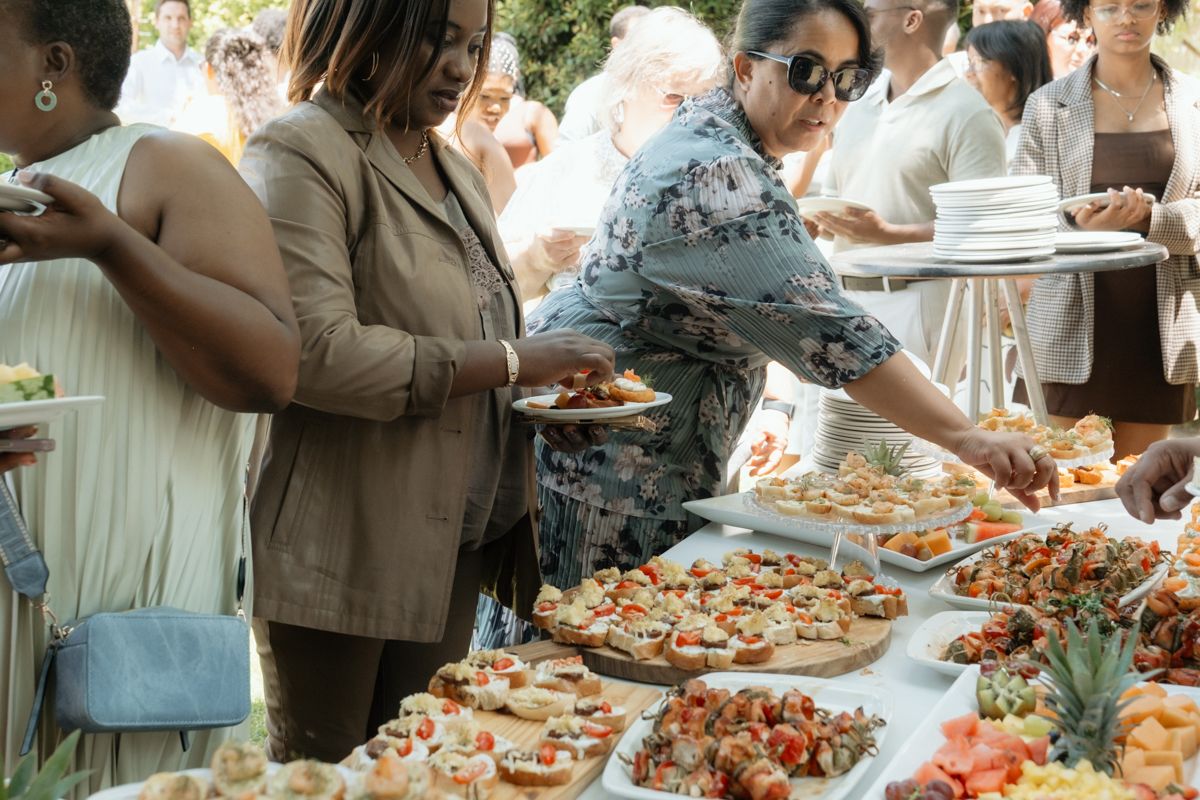 Guests serving themselves at an outdoor buffet table filled with colorful appetizers and finger foods at a catered event.