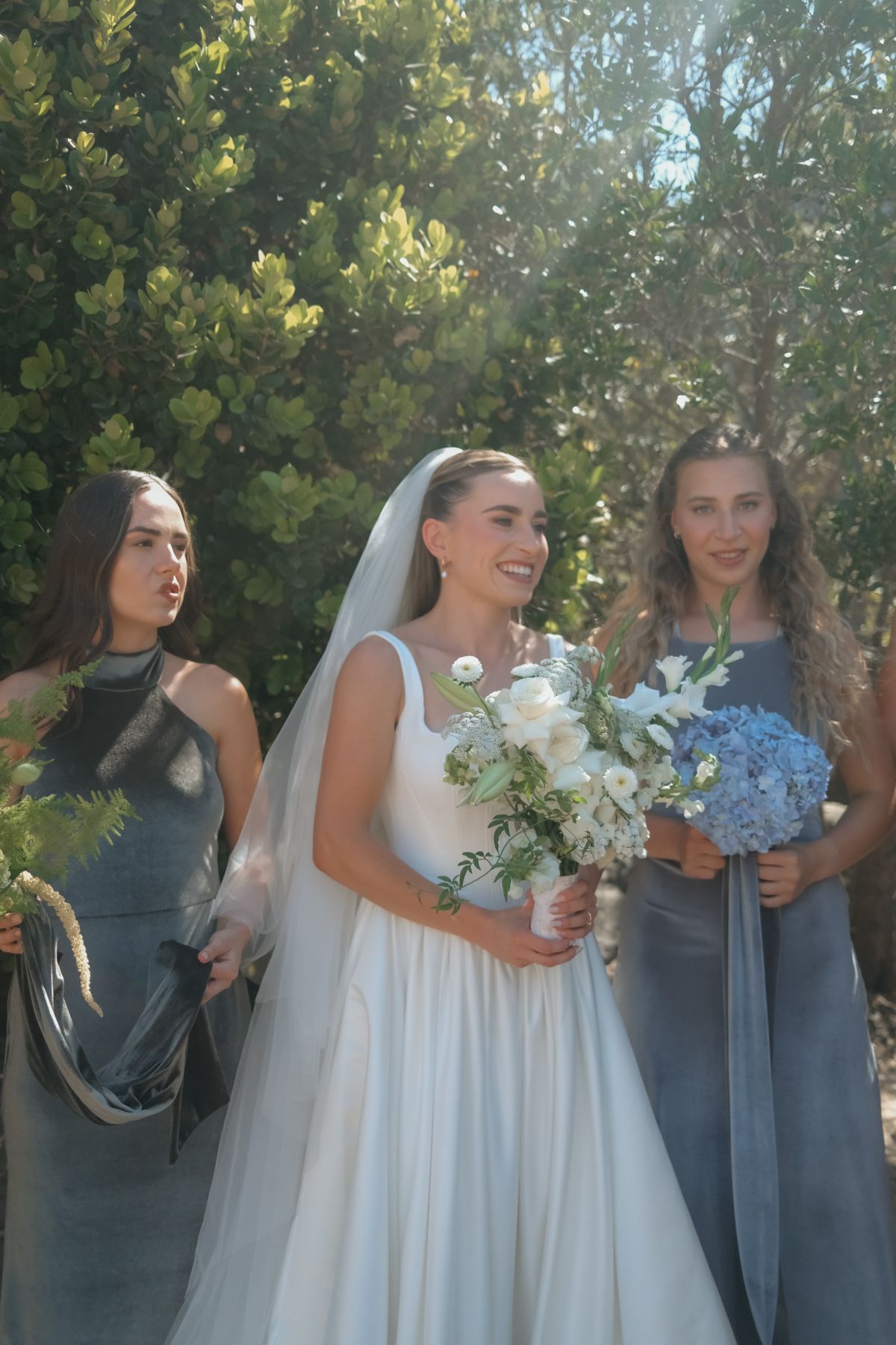 Smiling bride in white gown with veil holding bouquet flanked by two gray-dressed bridesmaids.