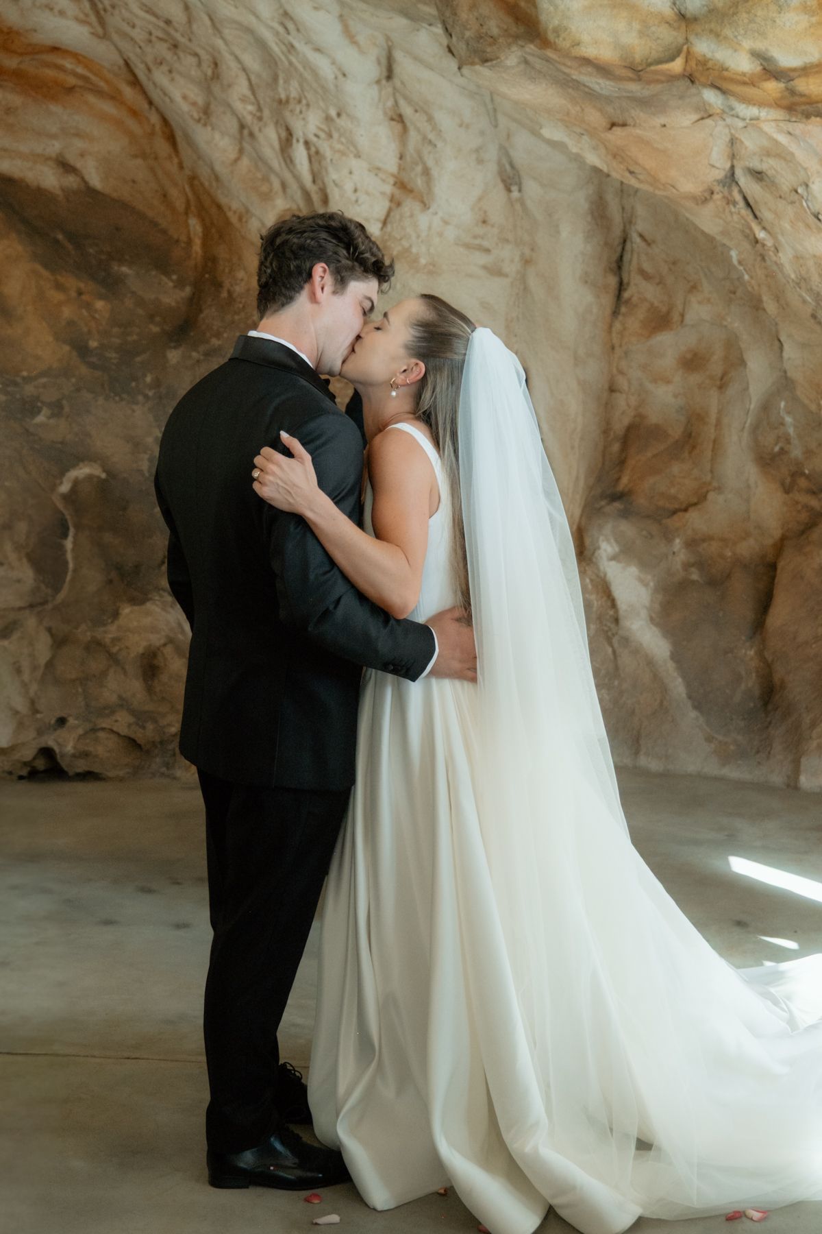 Bride and groom share first kiss in white gown and black tuxedo against a stunning natural stone wall backdrop.