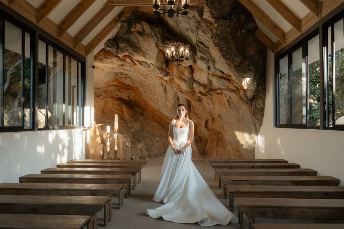 Bride in elegant white gown standing in sunlit chapel with natural rock wall backdrop.