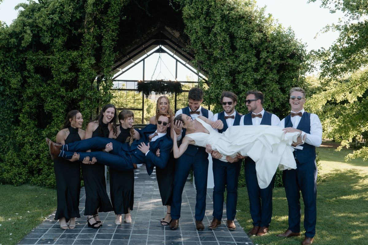 Wedding party in navy attire playfully carries bride along garden pathway under floral arbor.