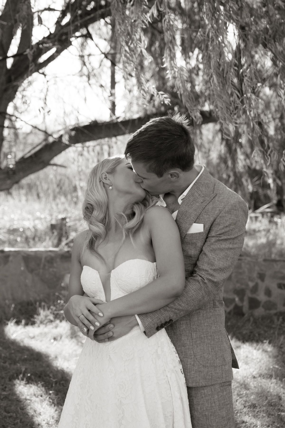Black and white photo of bride and groom sharing a romantic kiss under a willow tree on their wedding day.
