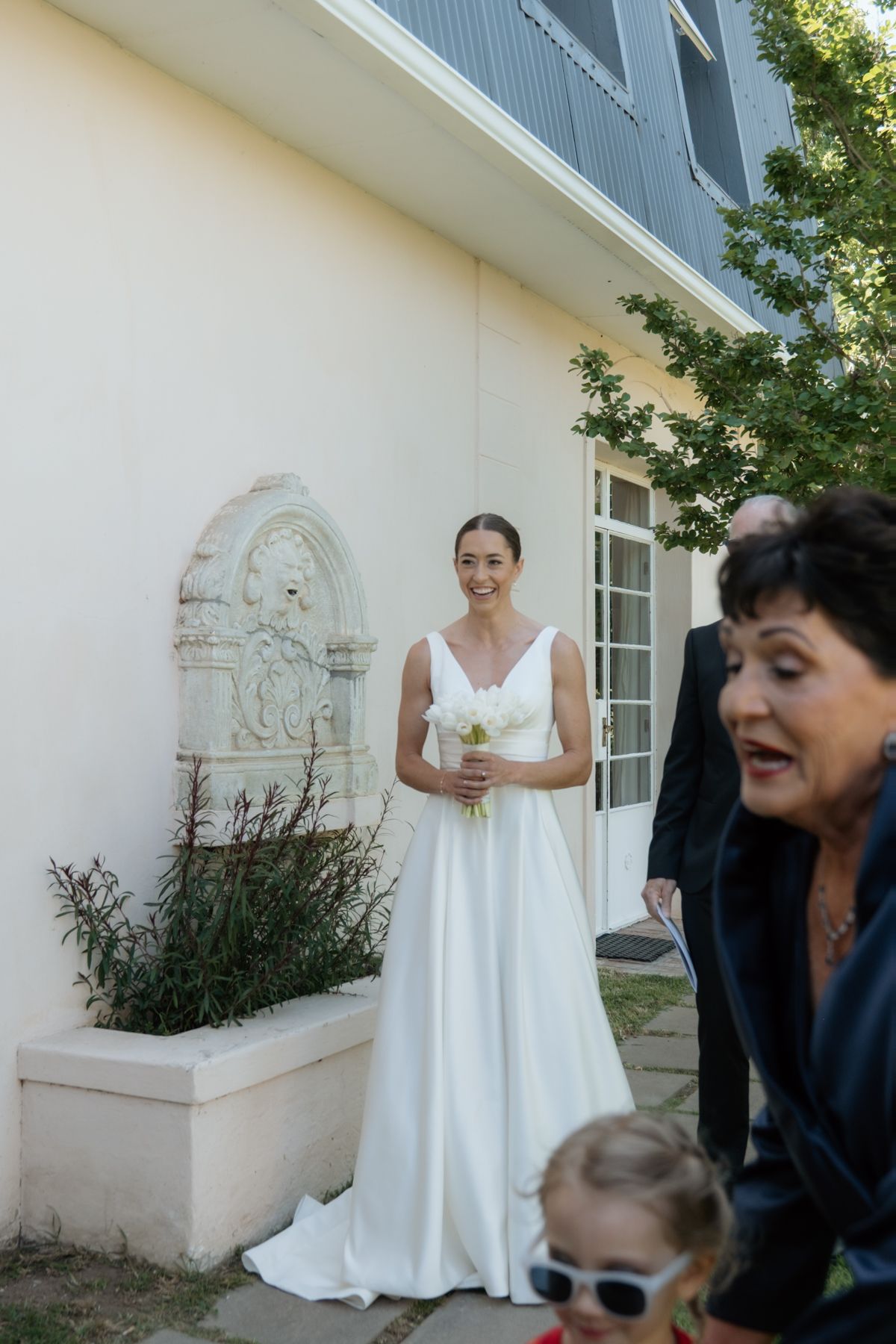 Smiling bride in a white gown holding a bouquet stands outside near a decorative wall fountain.