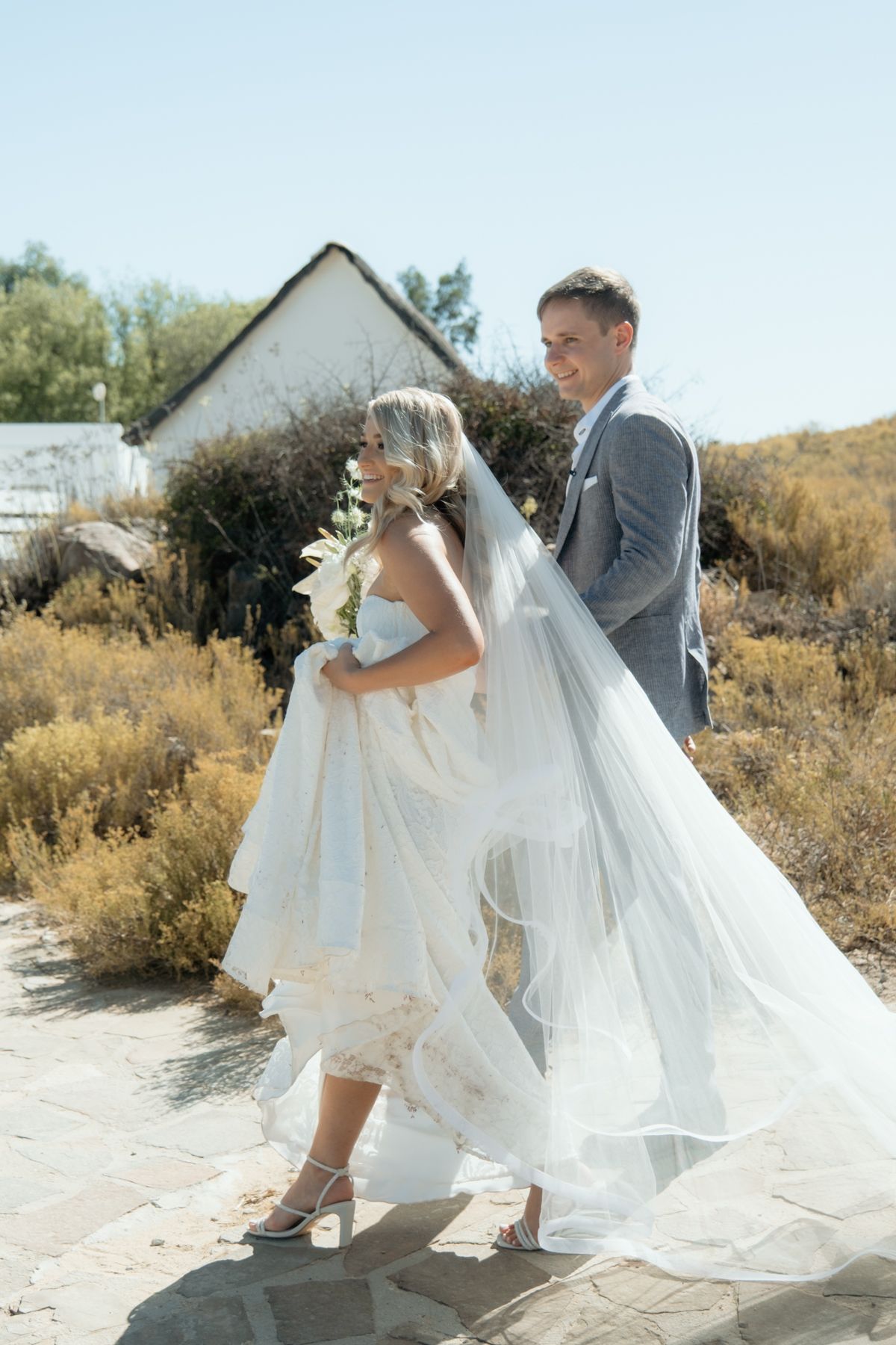 Bride and groom walking outdoors on a sunny day, bride wearing a lace gown with veil and holding bouquet.