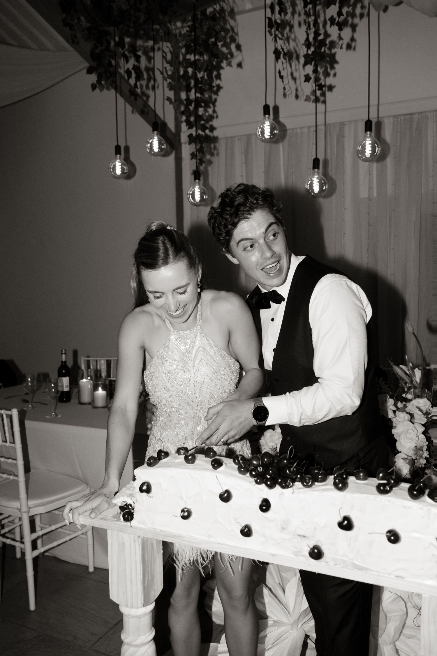 Black and white photo of a bride and groom joyfully cutting their wedding cake decorated with cherries.