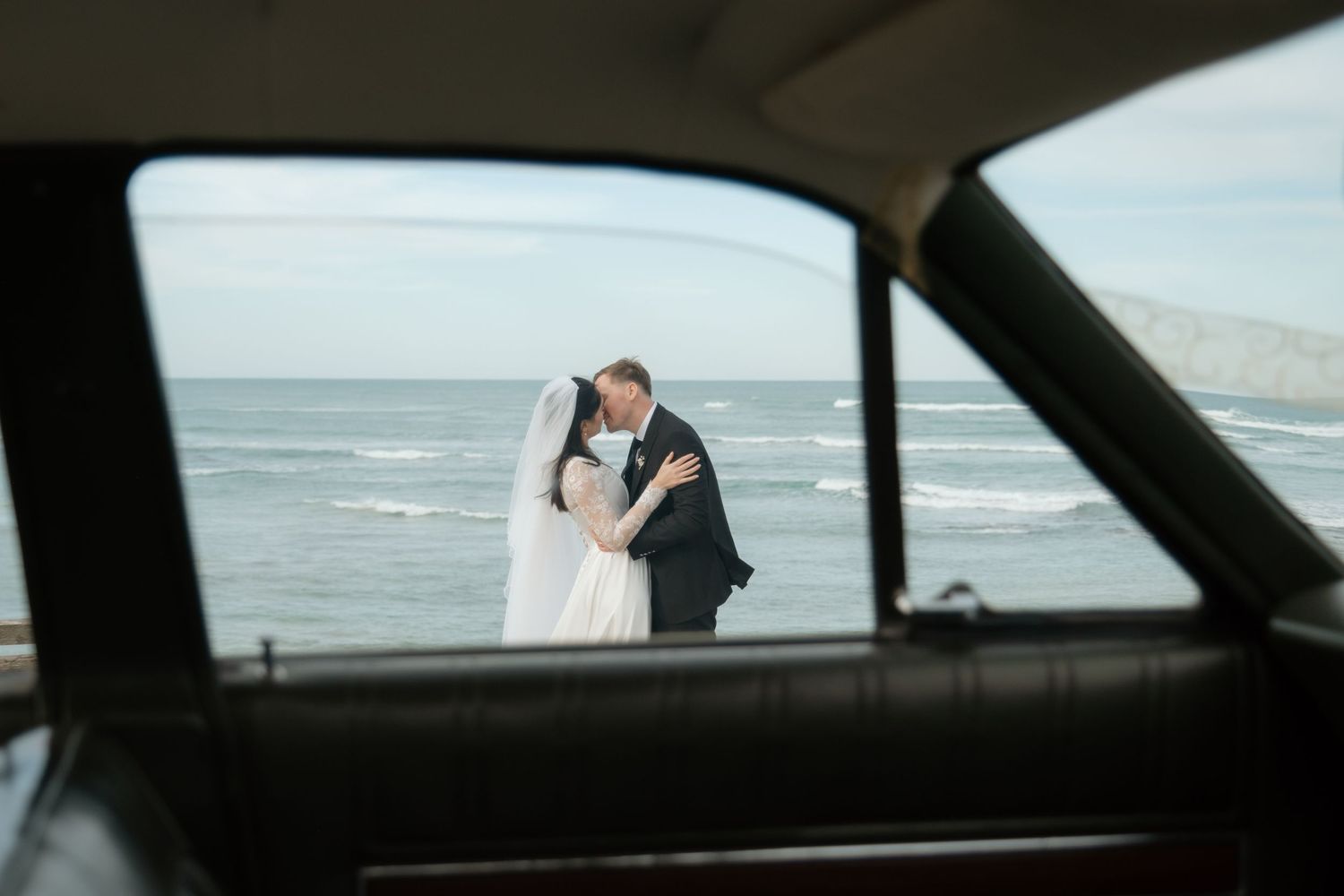 Bride and groom kissing by the ocean, framed through a vintage car window on their wedding day.