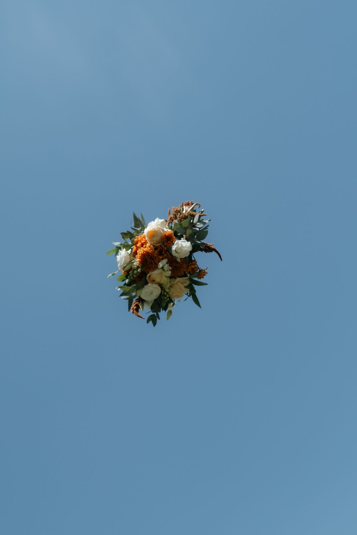 Wedding bouquet with white and orange flowers tossed against a clear blue sky during bouquet toss tradition.