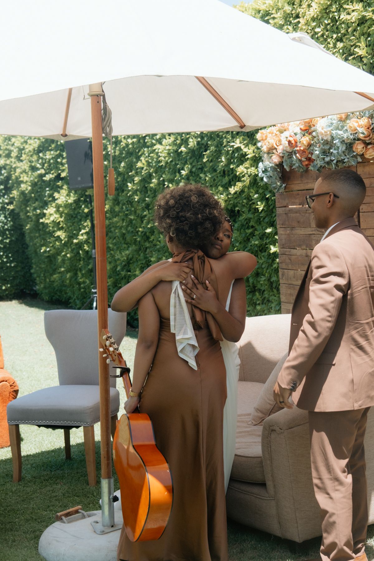 A woman in a brown gown holding a guitar embraces a man in a tan suit at an outdoor wedding celebration.