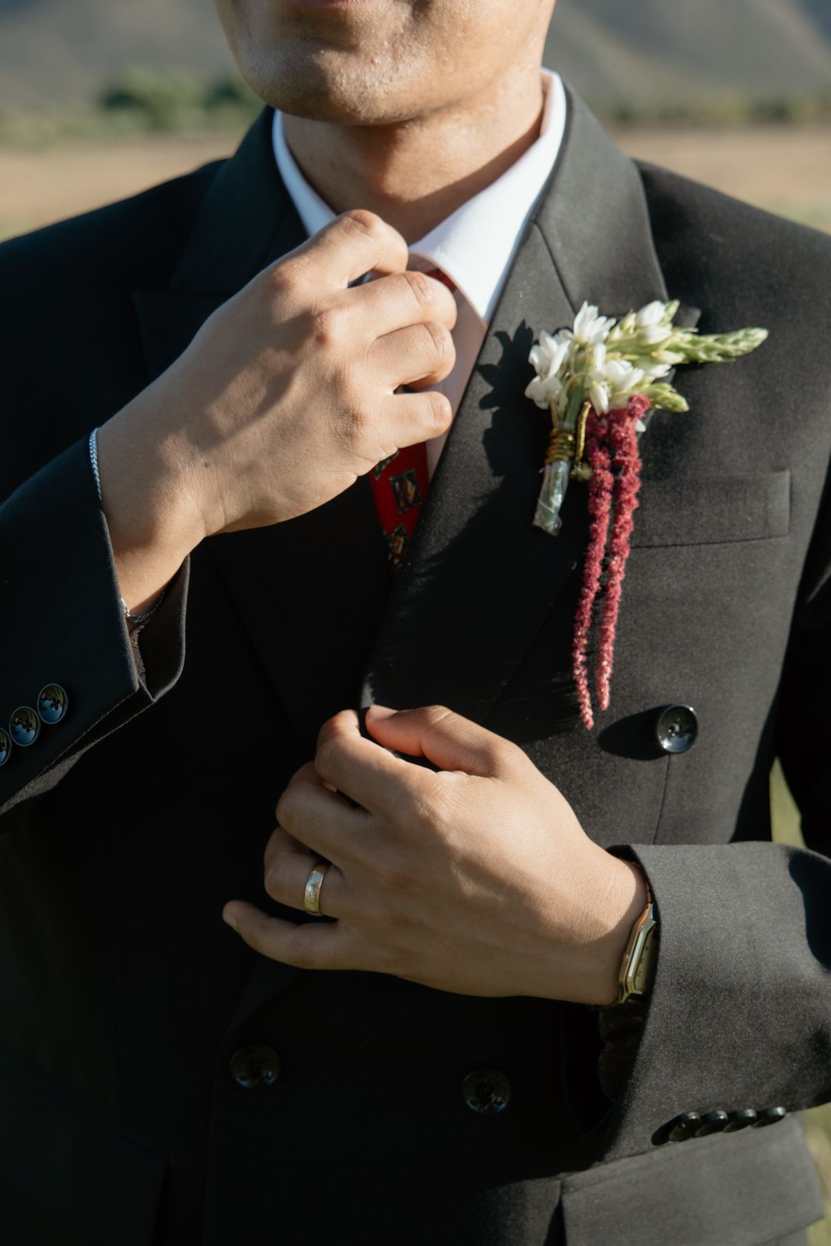 Close-up of groom in black suit adjusting tie, wearing wedding ring and floral boutonniere.
