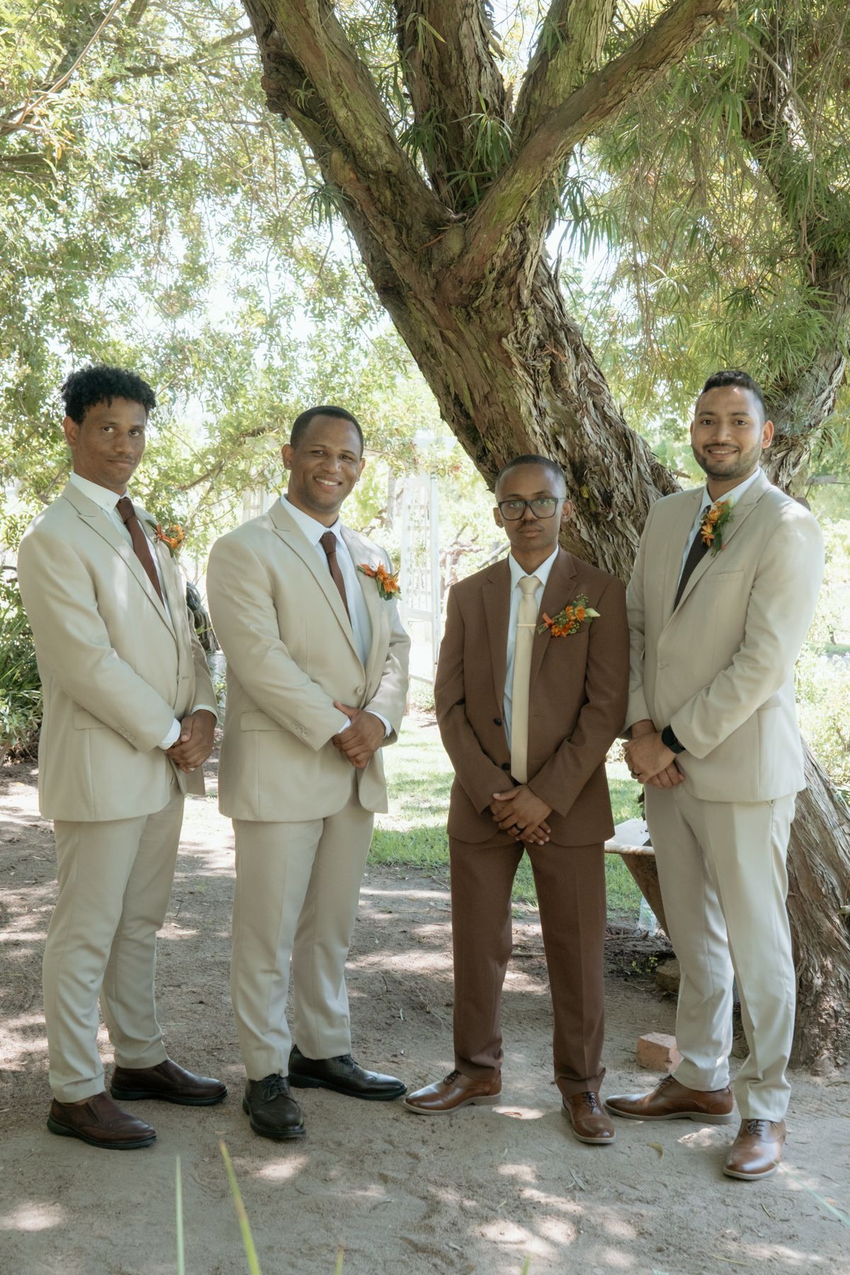 Four groomsmen wearing beige and brown suits with floral boutonnieres stand together under a tree at a wedding.