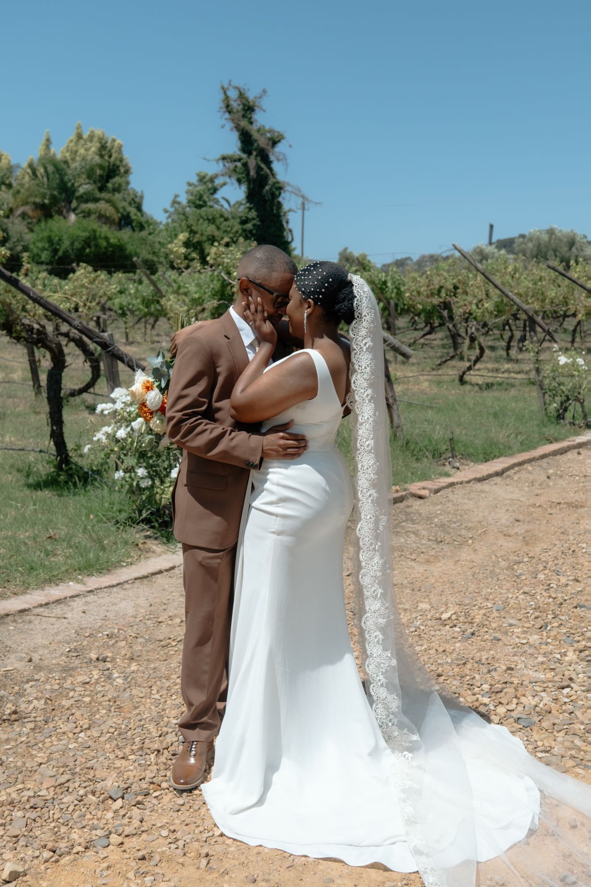 Black couple sharing a romantic kiss at outdoor wedding venue, bride in white gown with lace veil, groom in brown suit.