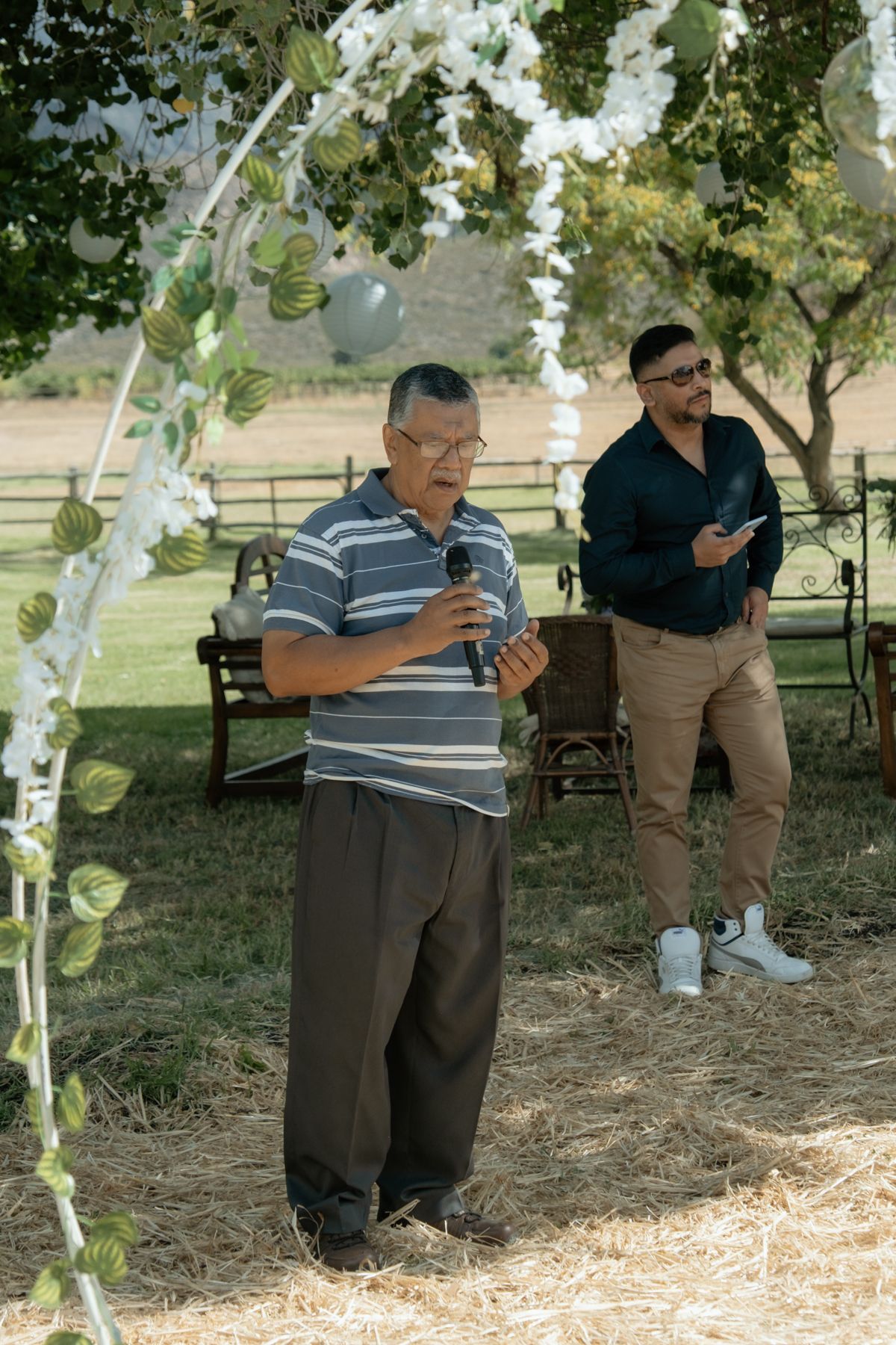Older man speaks into microphone at outdoor wedding ceremony beneath floral arch.