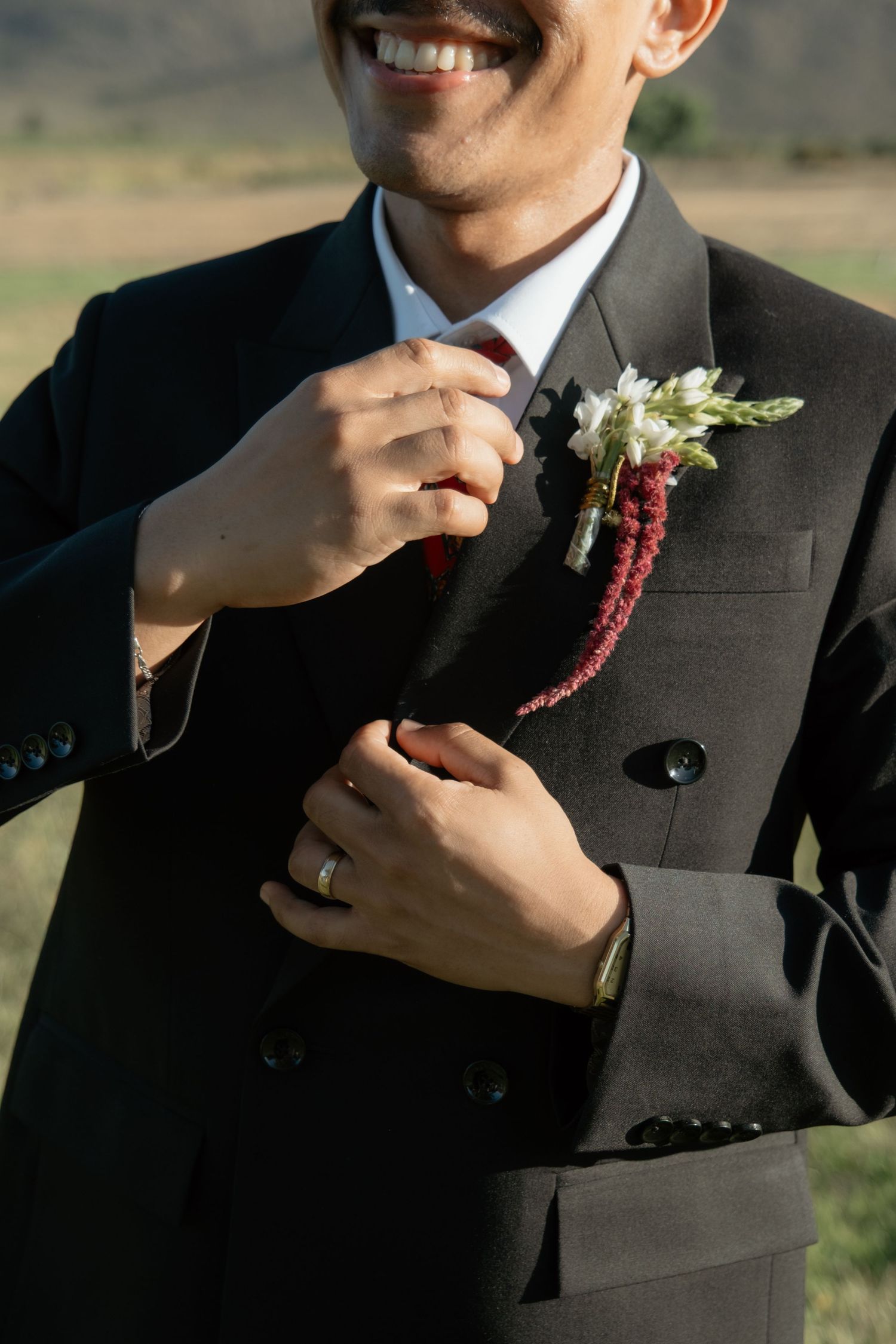 Smiling groom in black suit adjusting boutonniere with red and white flowers at outdoor wedding.