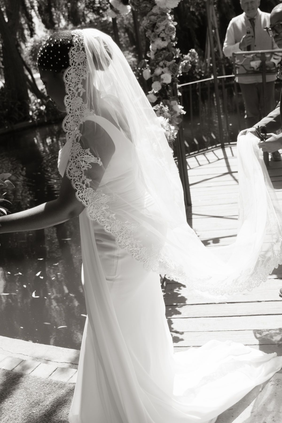 Black and white photo of bride in lace-trimmed veil and white gown at outdoor waterside ceremony.