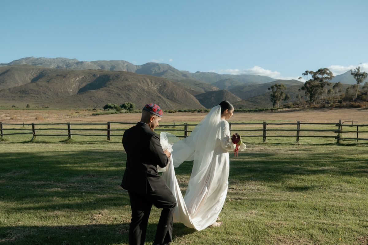Groom adjusts bride's veil in lush green field with stunning mountain backdrop.