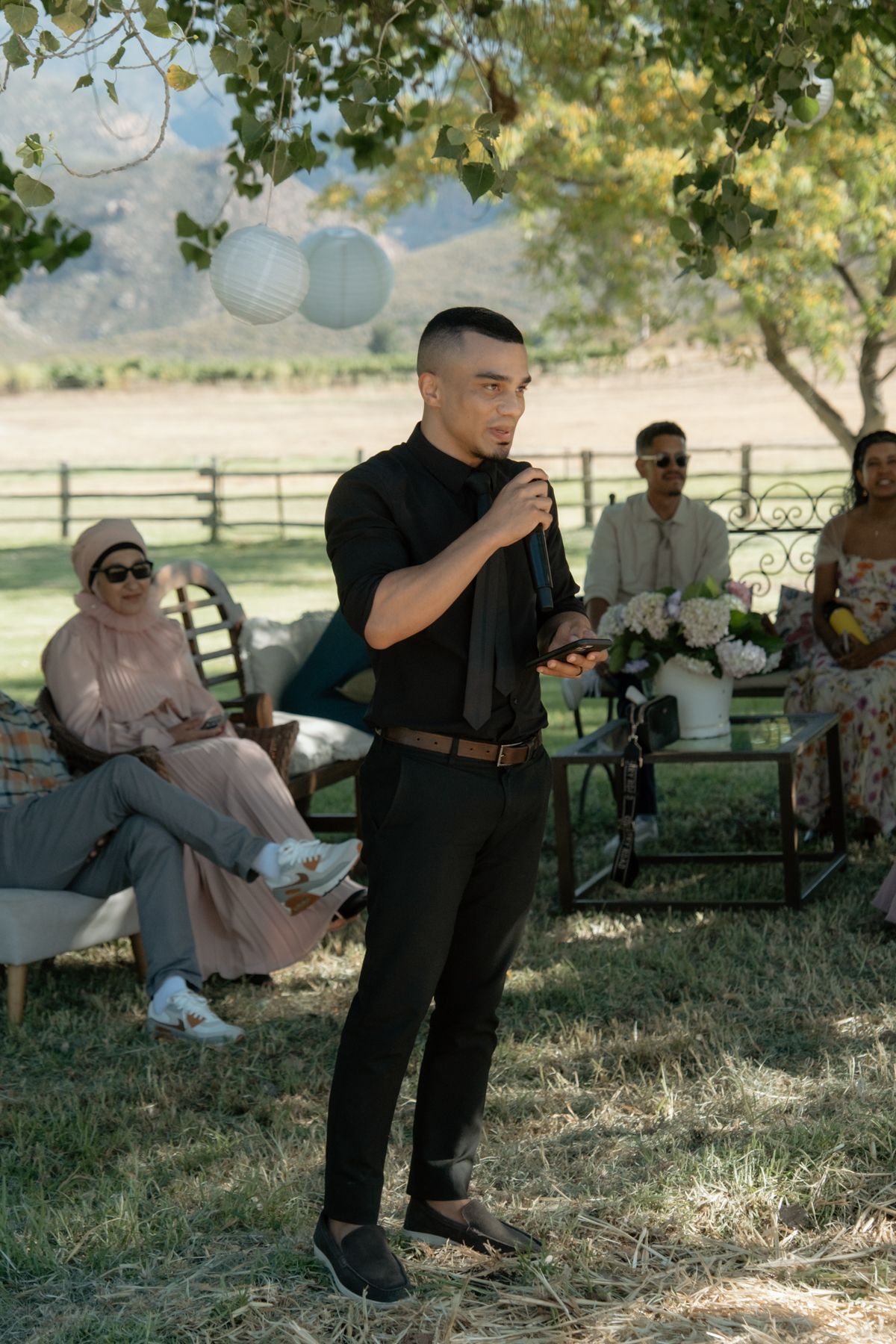Young man in black outfit giving a speech at an outdoor wedding reception under a tree.