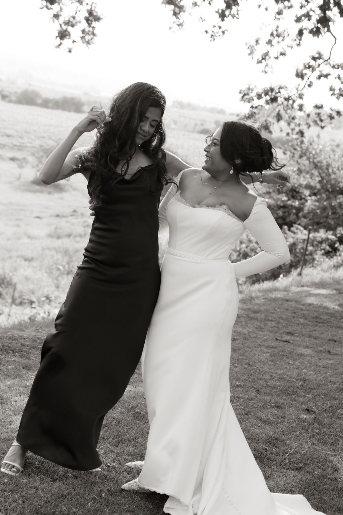 A bride in a white gown and bridesmaid in black dress dance joyfully outdoors in black and white.