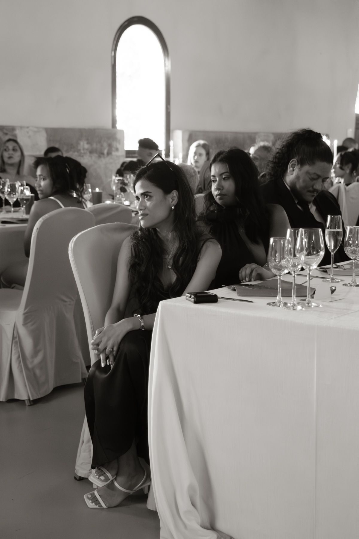 Black and white photo of elegantly dressed wedding reception guests seated at tables with wine glasses.