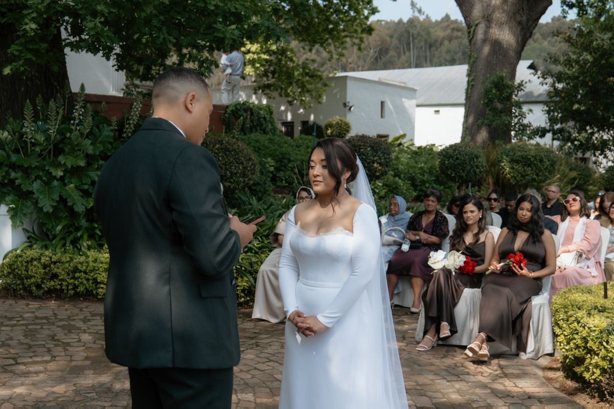 Bride in elegant white gown exchanges vows with groom during intimate outdoor wedding ceremony with guests seated.