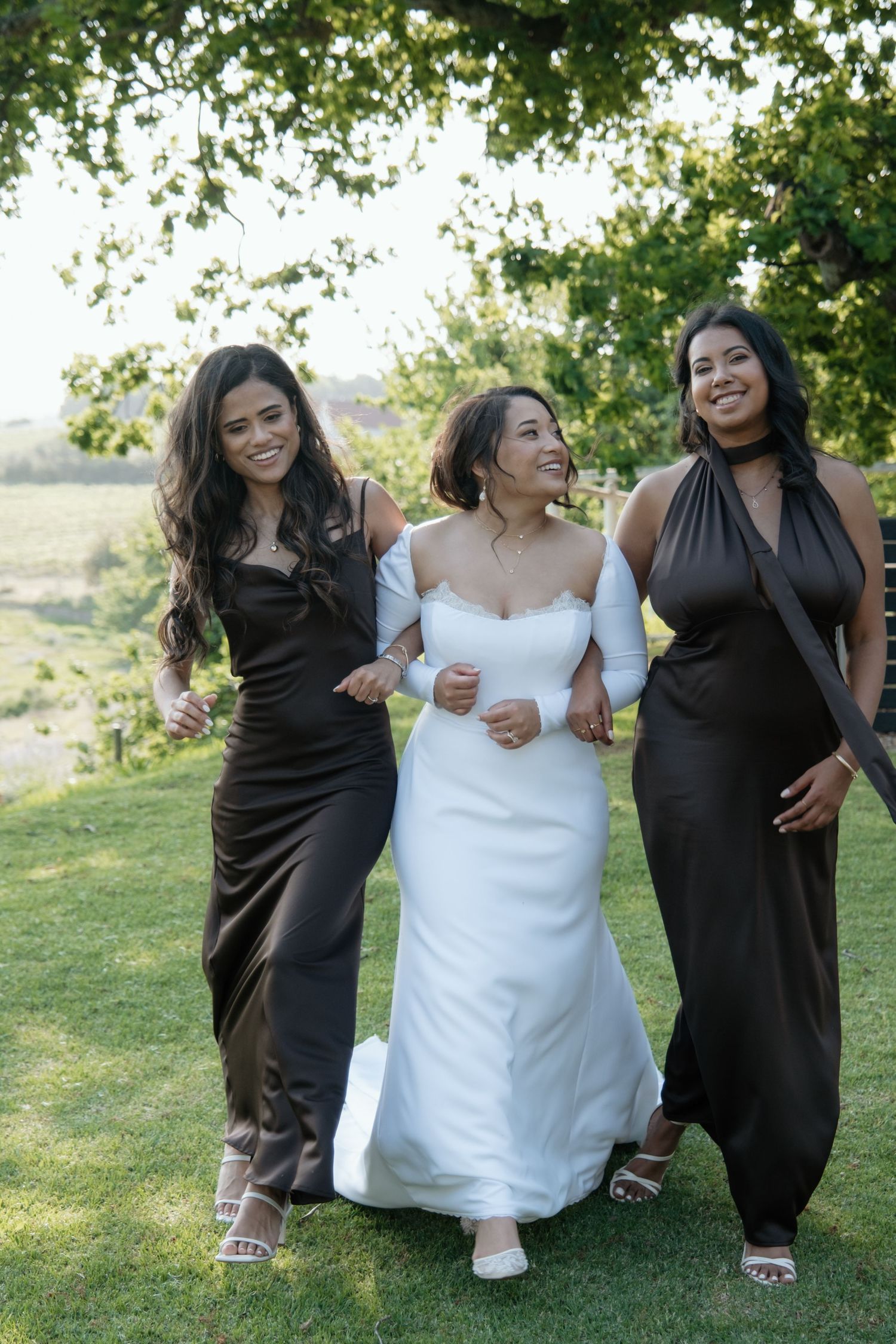 Bride in white strapless gown walking arm in arm with two bridesmaids in dark satin dresses outdoors.