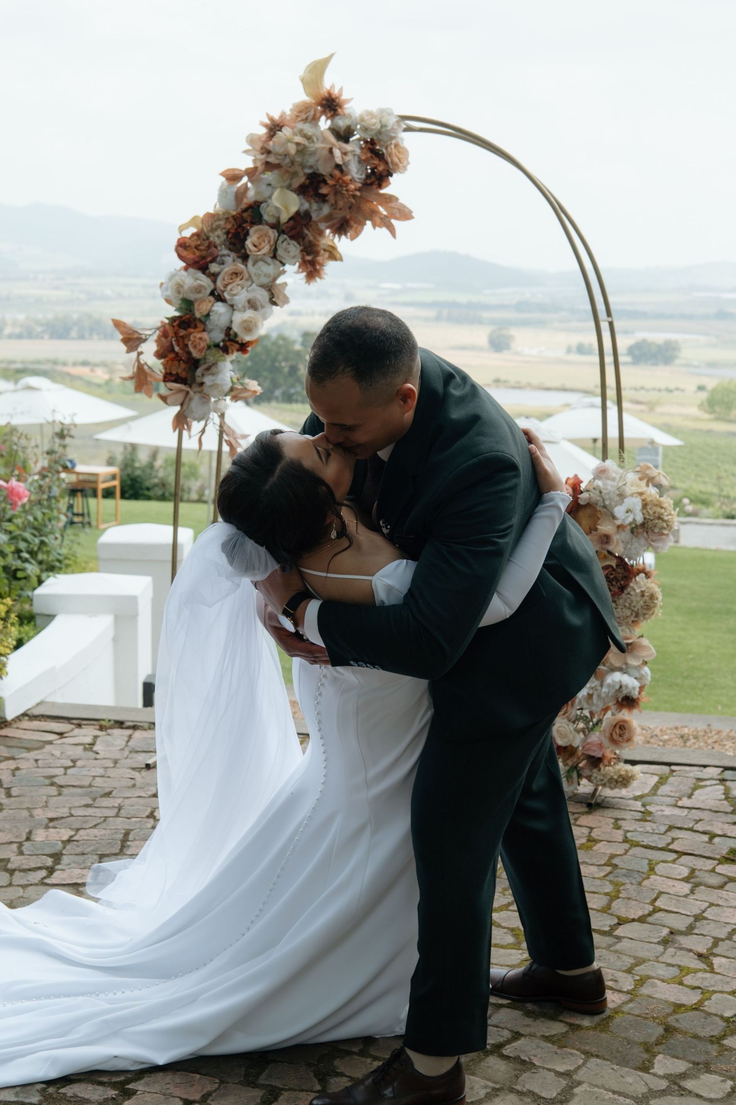 Bride and groom share first kiss under floral arch at outdoor wedding ceremony.