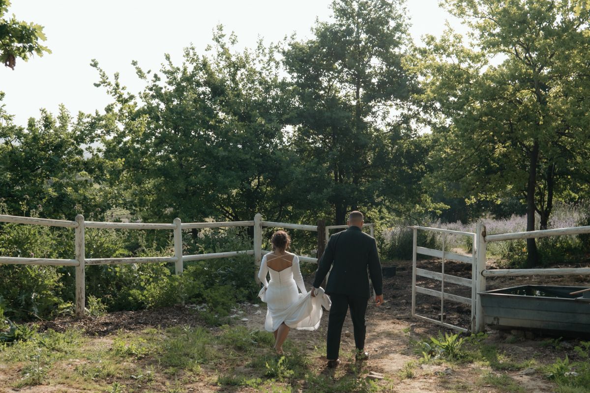 Bride and groom holding hands walking toward a wooden fence surrounded by lush green trees.