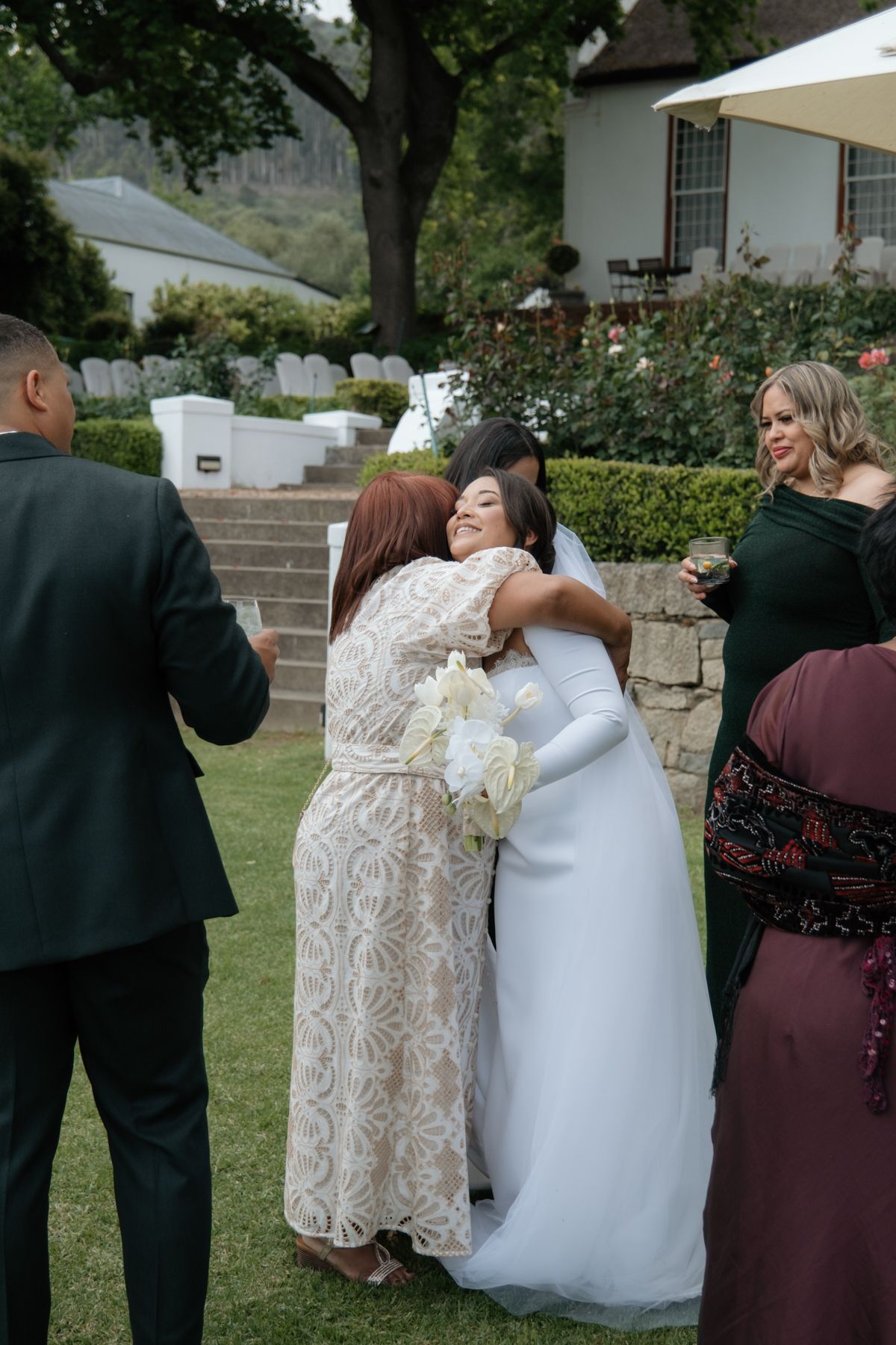 A bride in a white gown embraces a guest in a lace dress at an outdoor wedding reception.