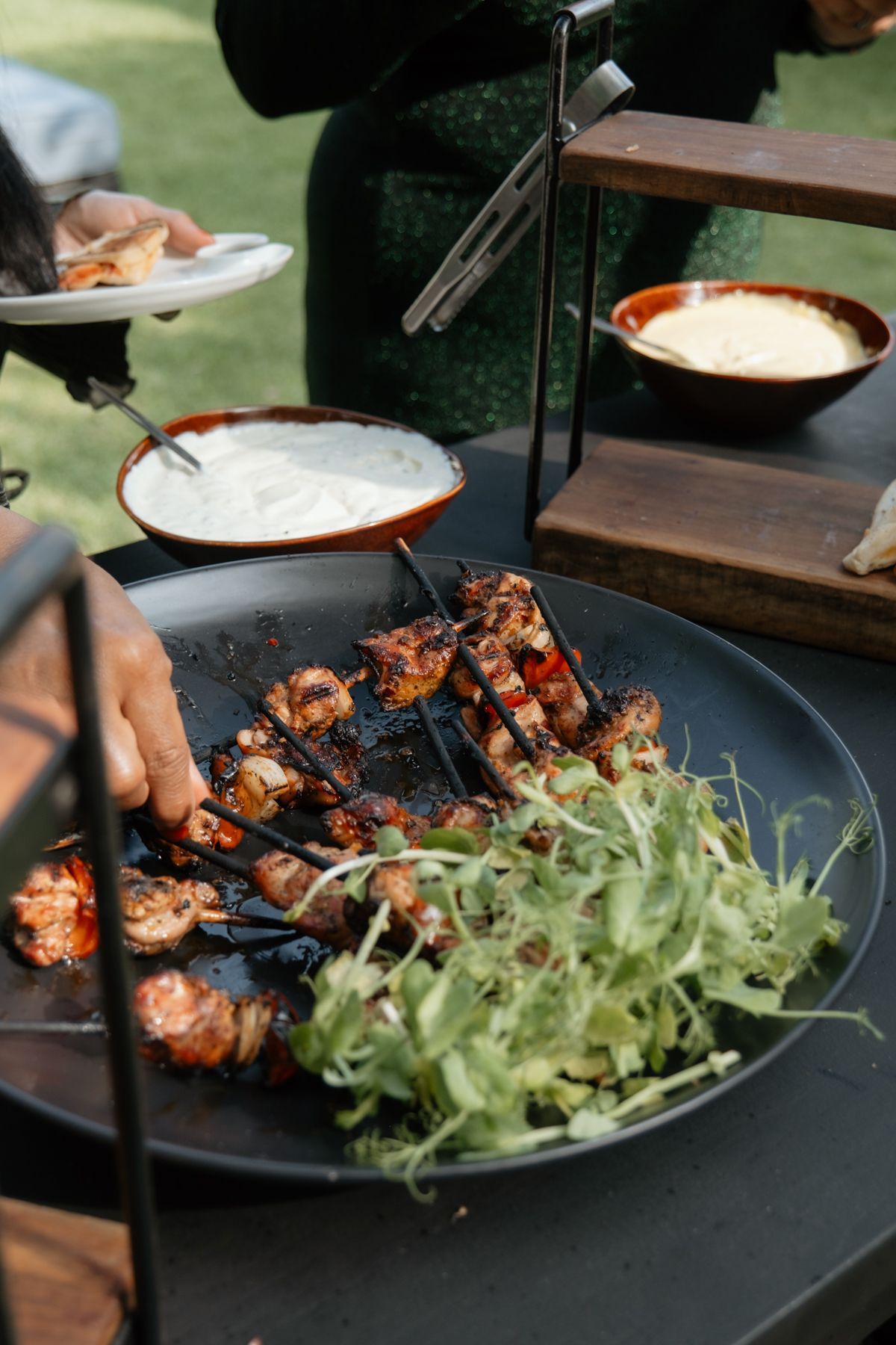 Grilled meat skewers with fresh greens served on large black platter at outdoor wedding reception.
