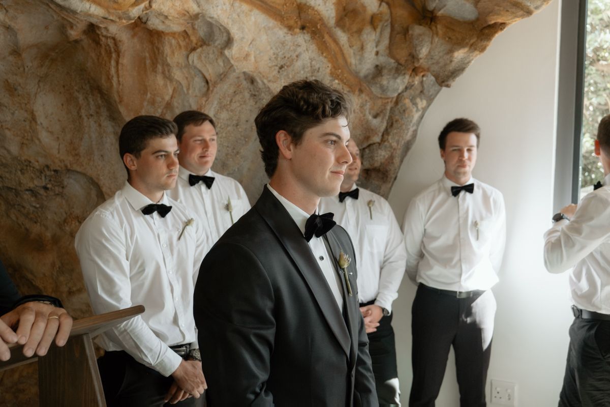 Groom in black tuxedo stands with groomsmen in white shirts and bow ties at a wedding venue.