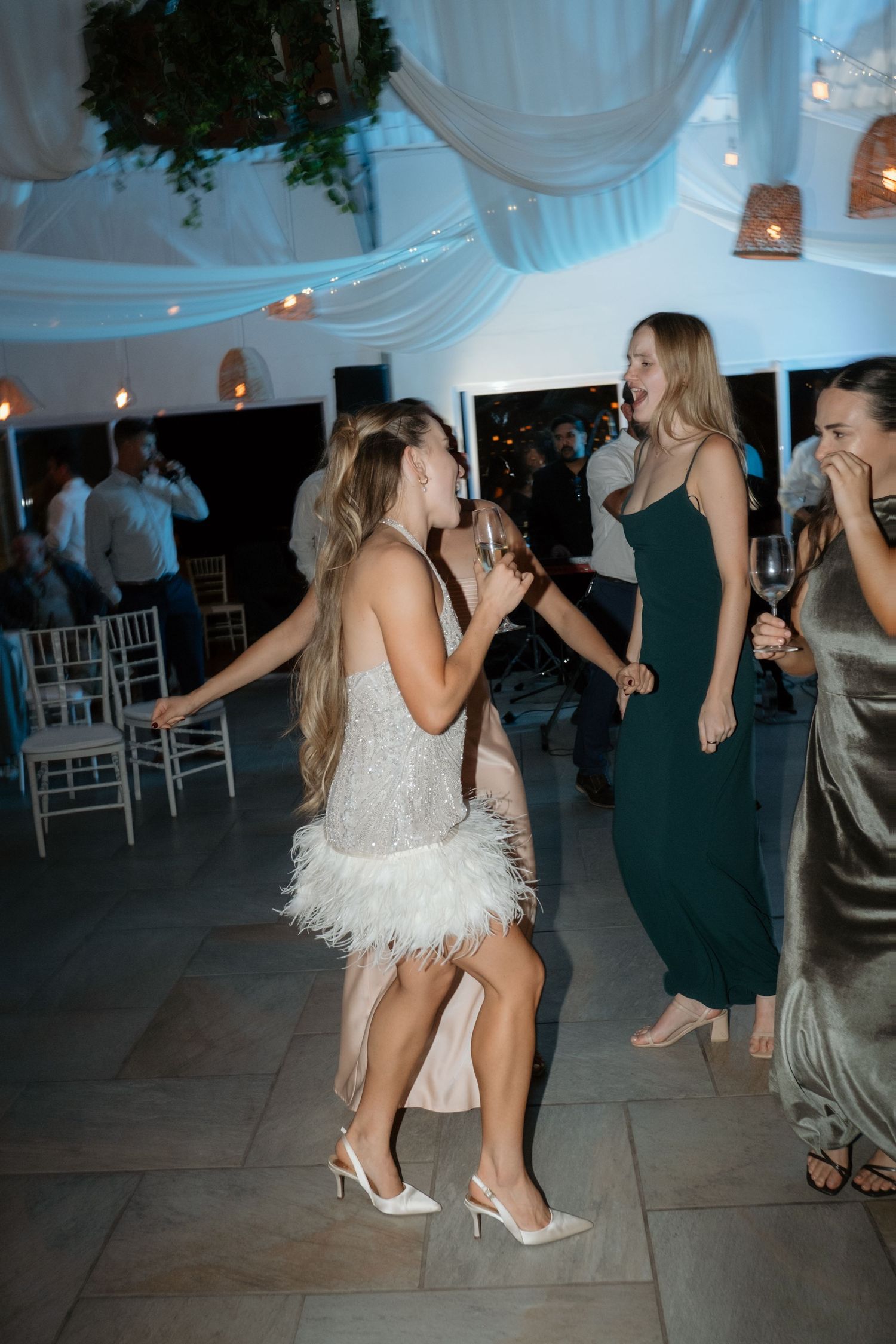 Two women dancing joyfully at a wedding reception, one in a white feathered dress and another in an emerald green gown.