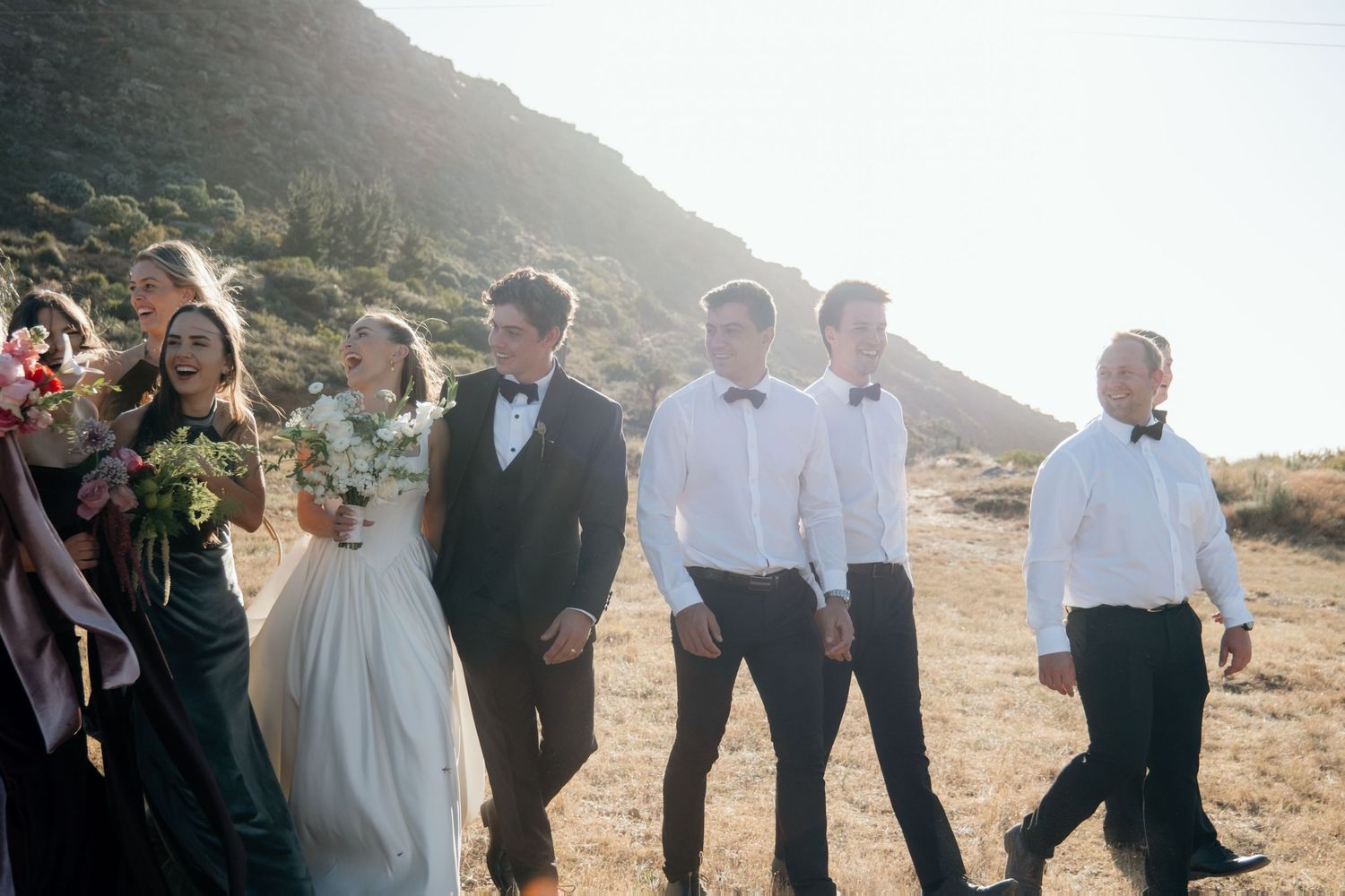 Wedding party in formal attire walking together outdoors with mountain backdrop in golden sunlight.