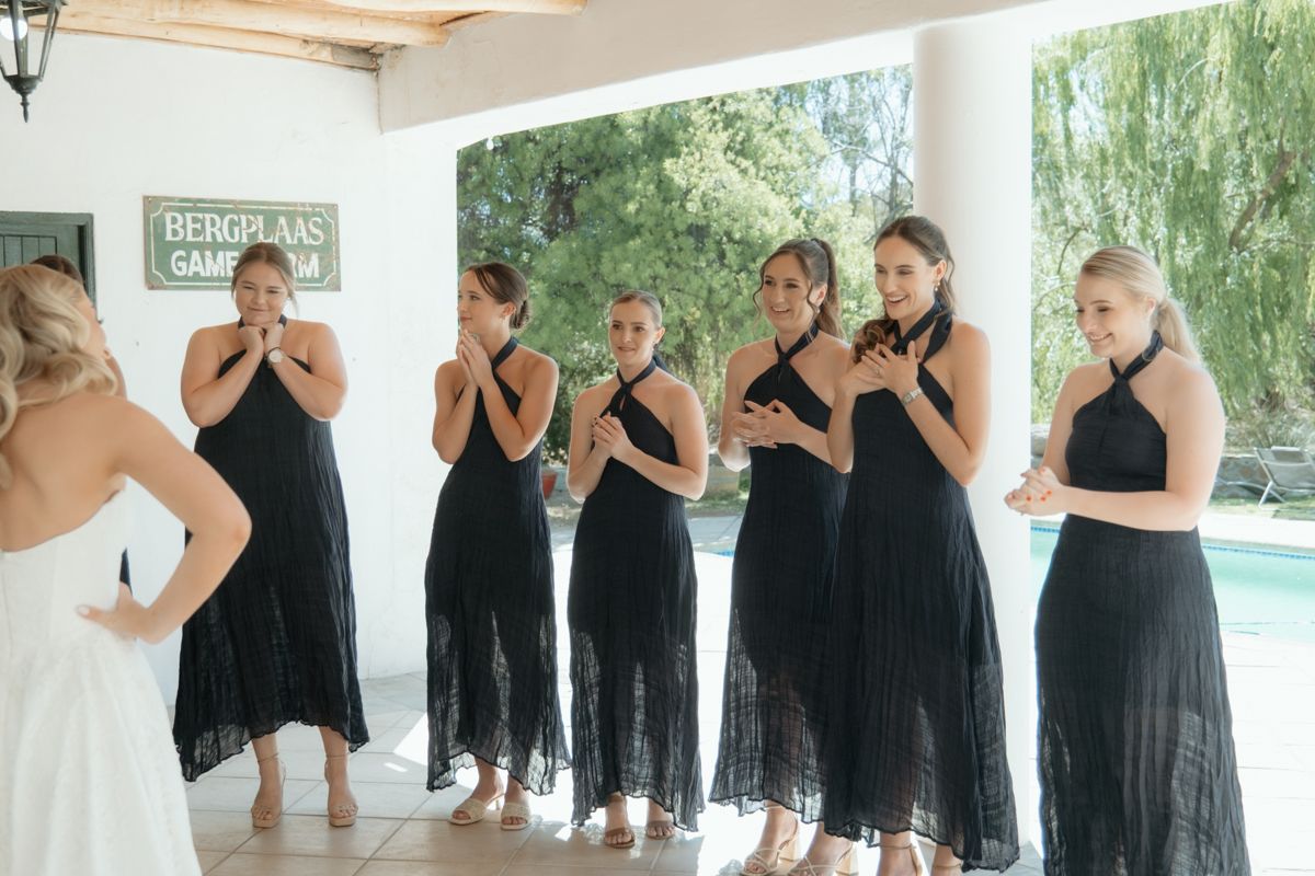 Bridesmaids in black dresses react with joy as bride reveals her wedding gown poolside.